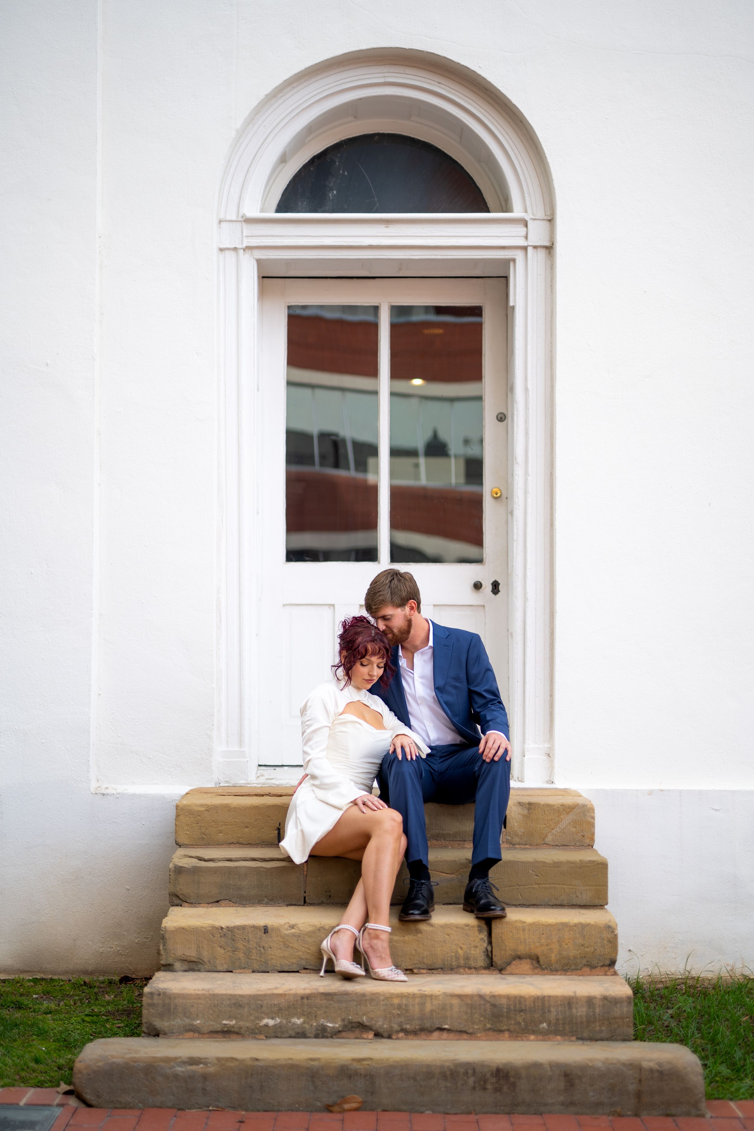 A couple sits intimately on stone steps in front of a white building with an arched doorway.