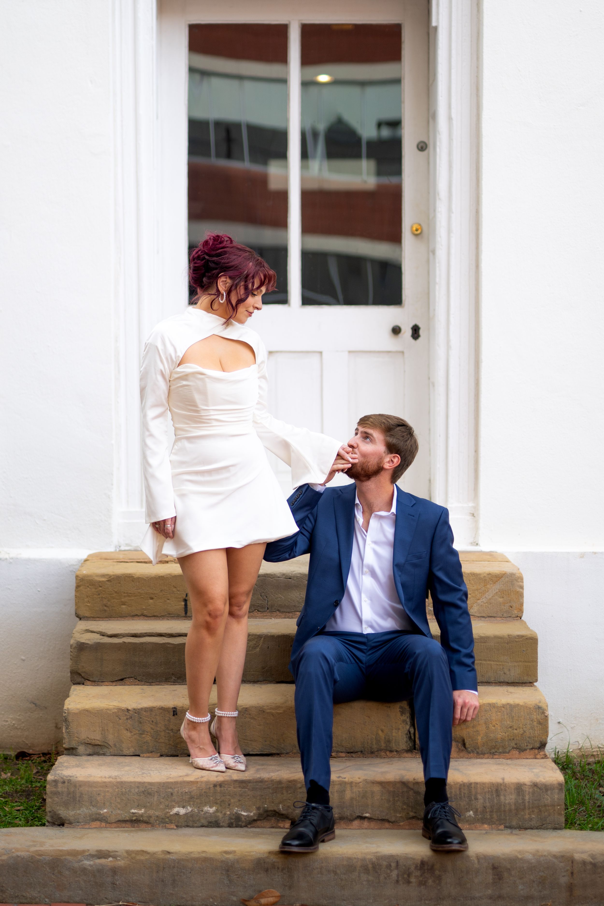 A romantic couple in contrasting outfits poses on white stone steps in front of a classic doorway.