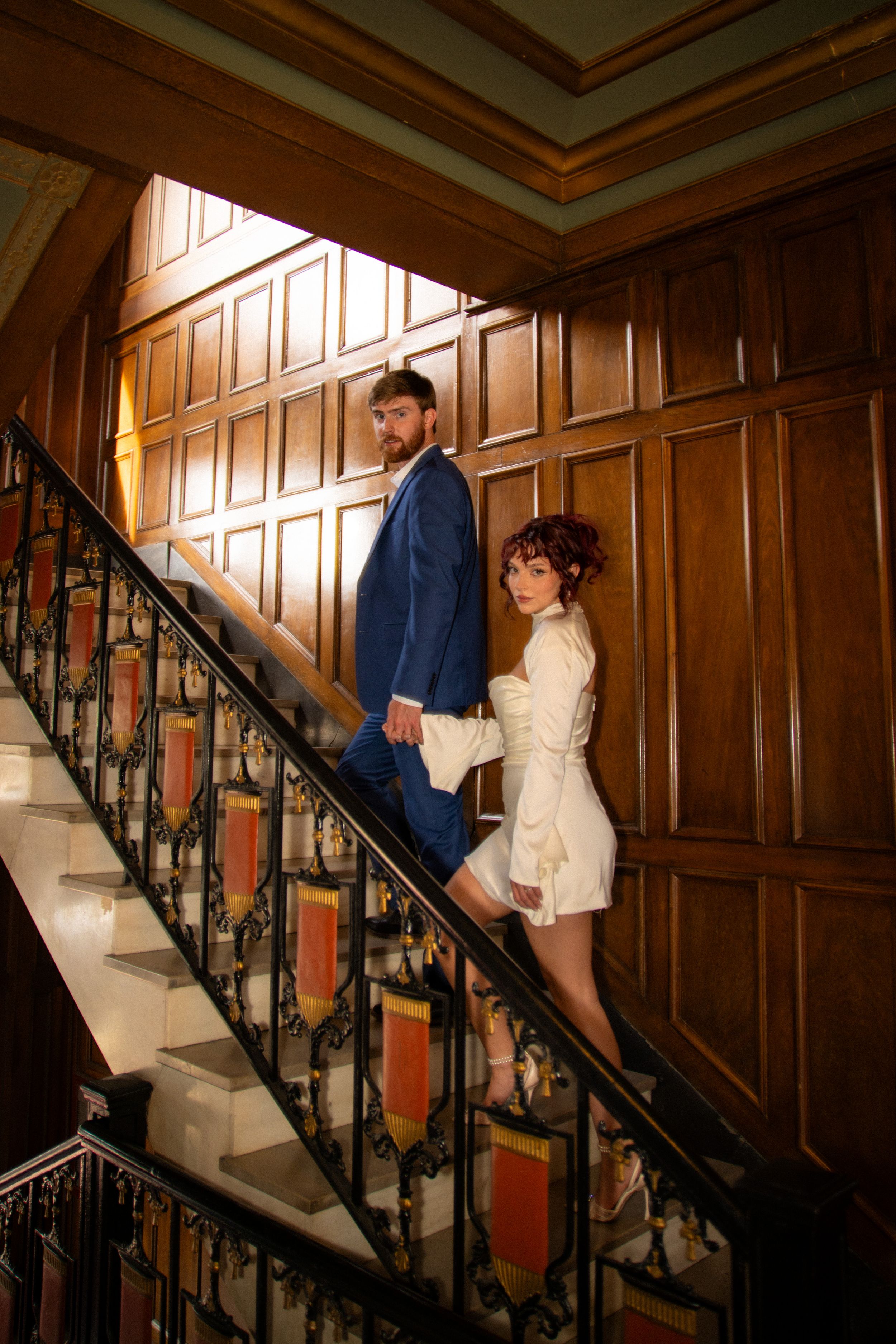 Couple descending a grand wooden staircase with ornate details and iron railings.
