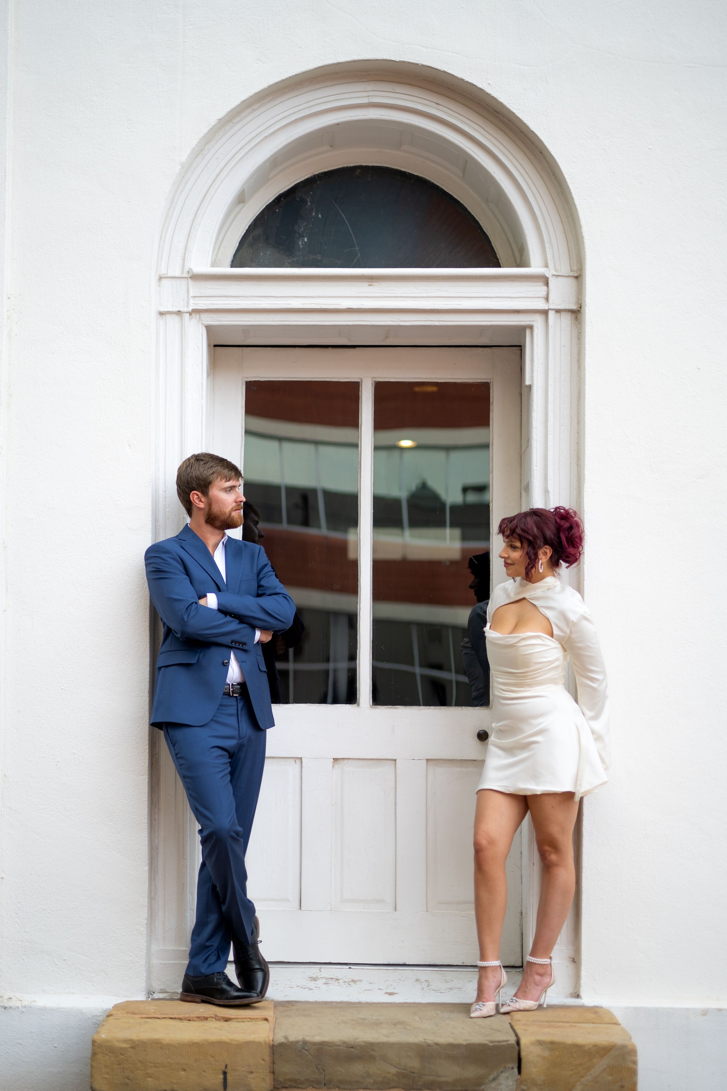 Playful couple shares a fun moment against a white building with arched windows.