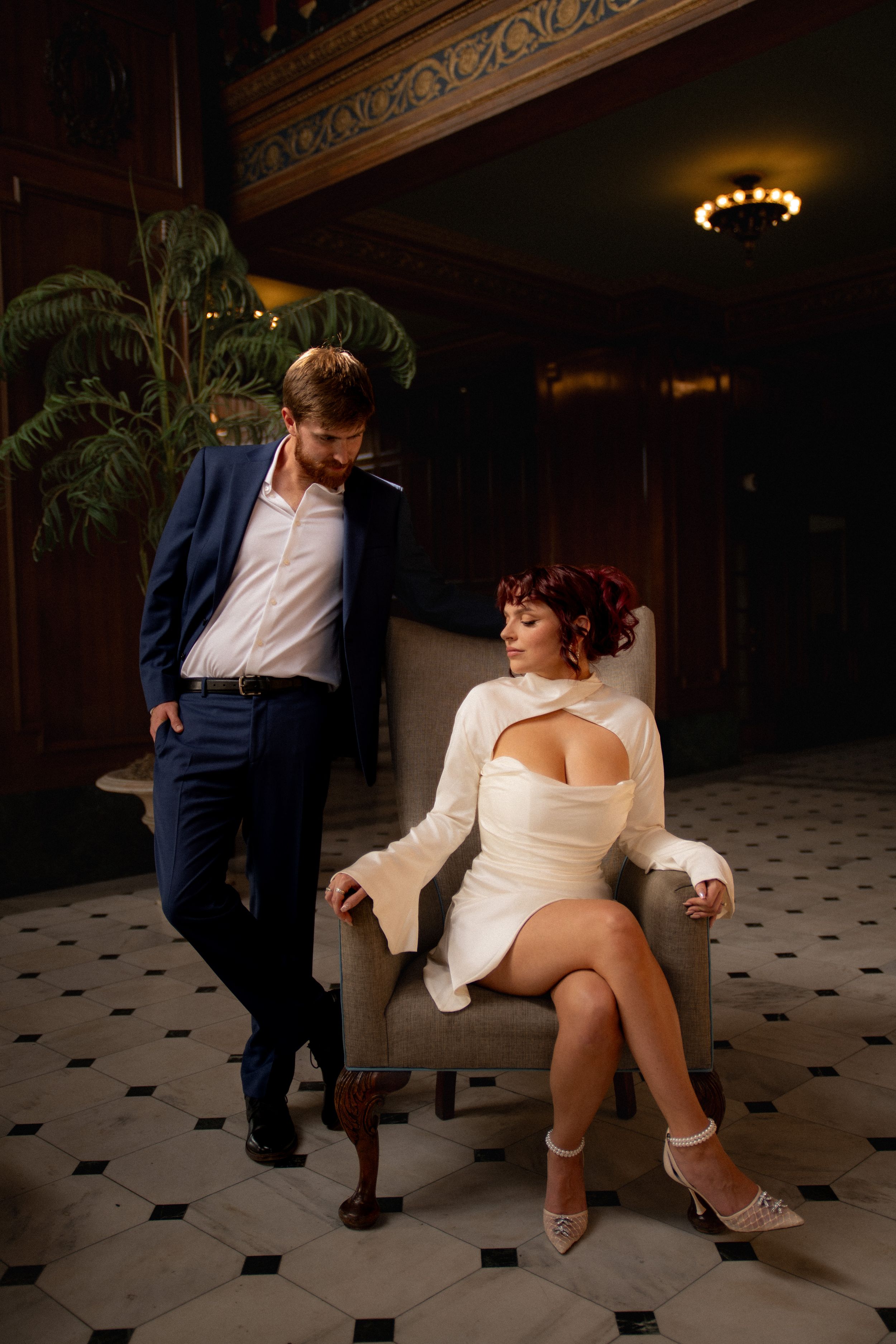 A glamorous couple poses in formal attire inside an elegant venue with ornate details and mood lighting.