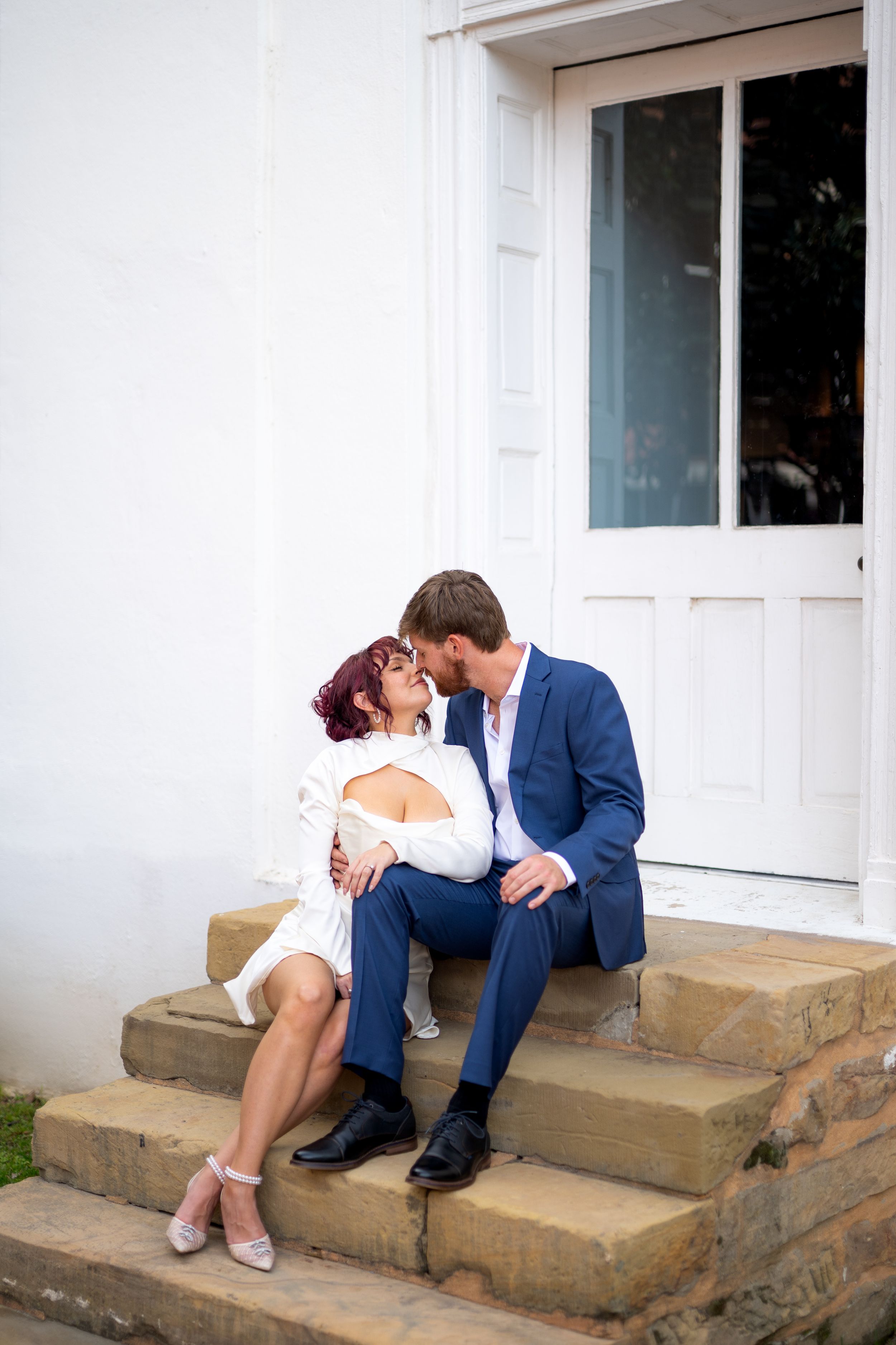 Romantic couple embraces on stone steps in front of a white colonial-style doorway.