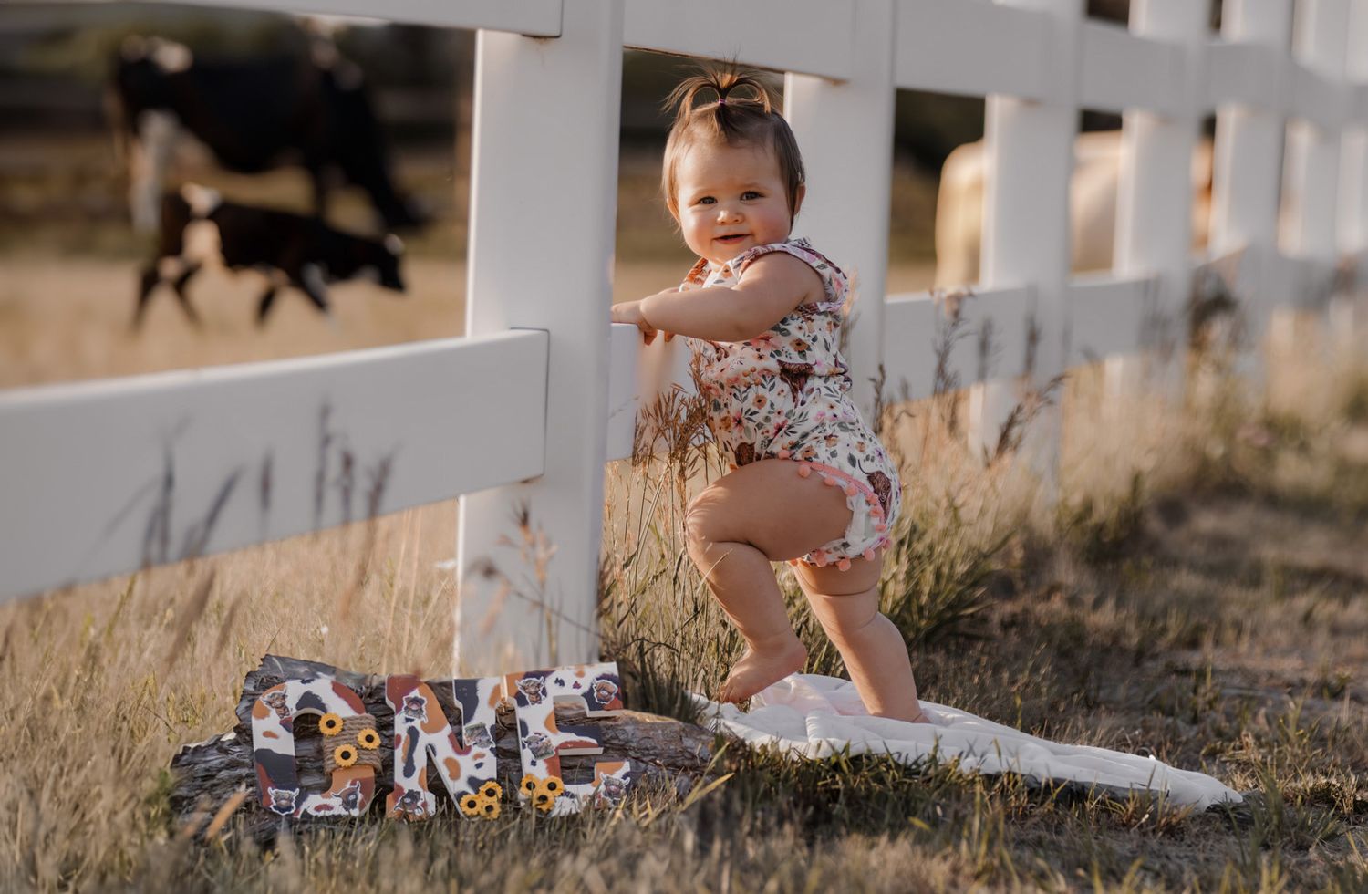 Small child in floral outfit plays near white fence during golden hour.