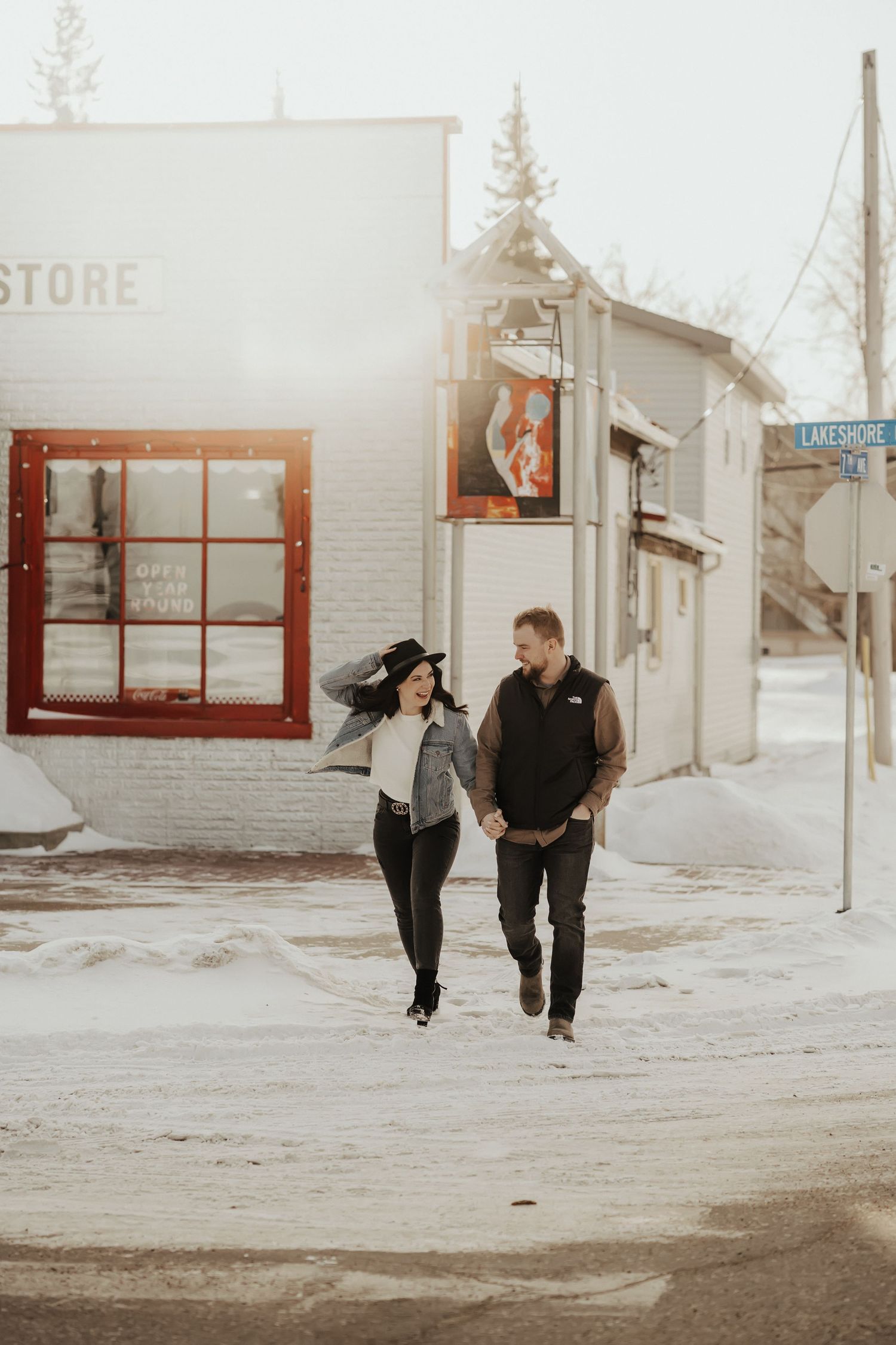 Couple walking together in a snowy small town street with rustic storefronts in the background.