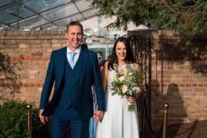 A newlywed couple poses in front of a rustic brick wall in formal wedding attire.