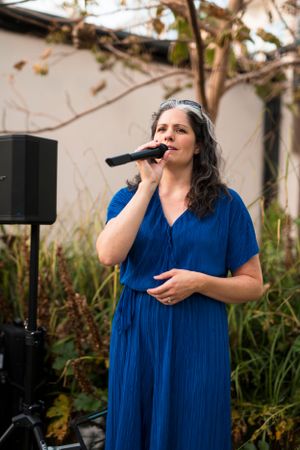 A performer in a blue dress sings into a microphone during an outdoor event.