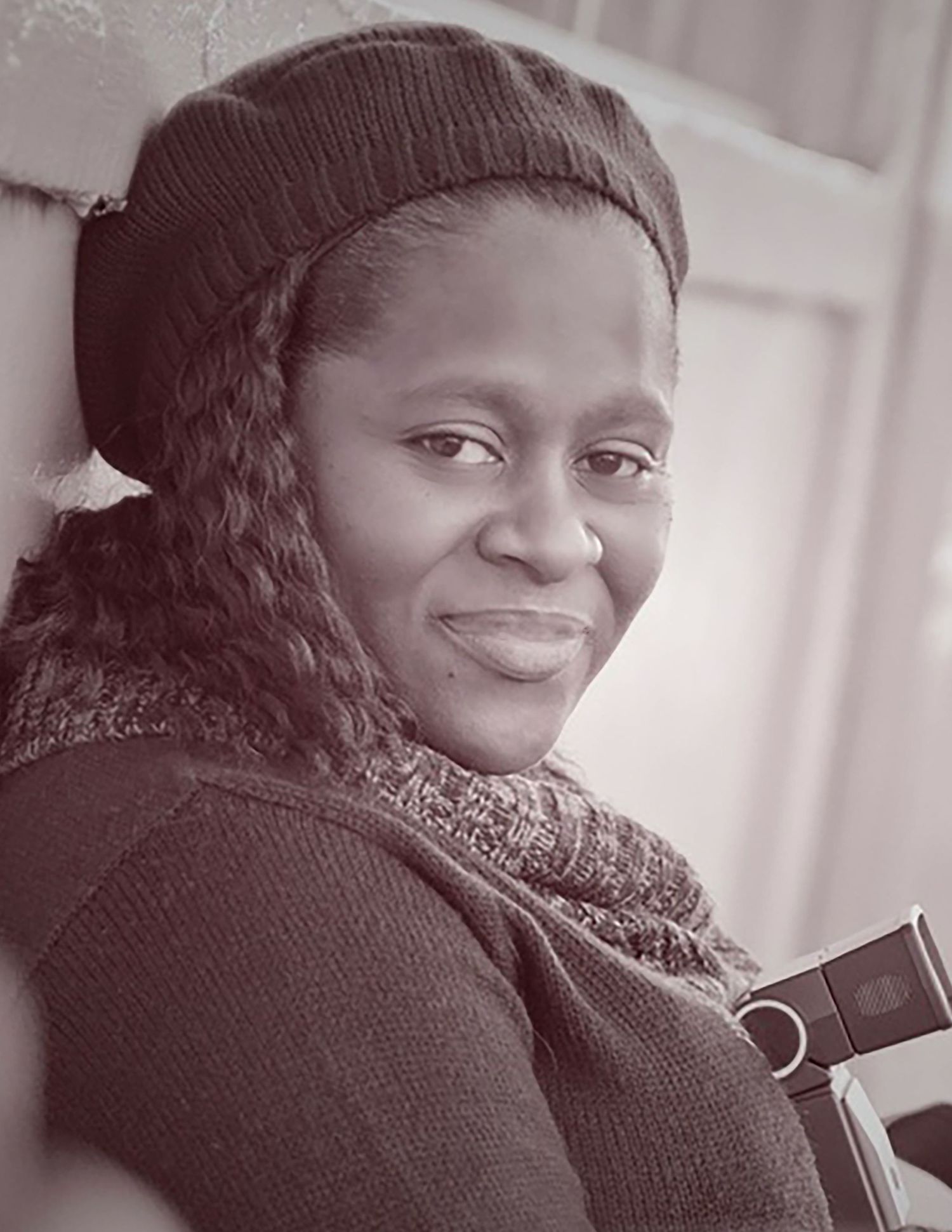 A portrait photo in black and white of someone wearing a knit cap and sweater while smiling warmly at the camera.