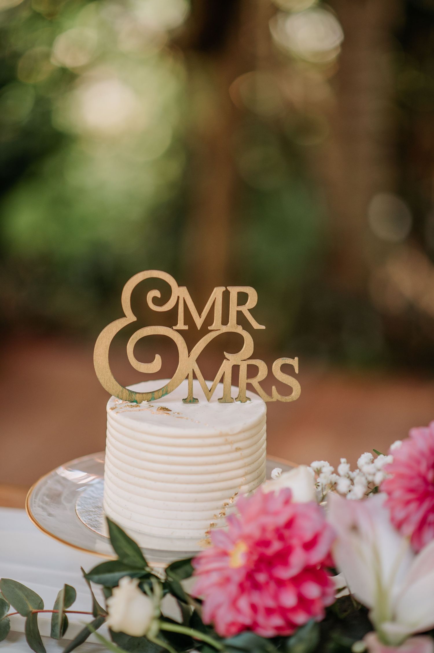 White wedding cake decorated with gold 'Mr & Mrs' topper and pink floral accents.