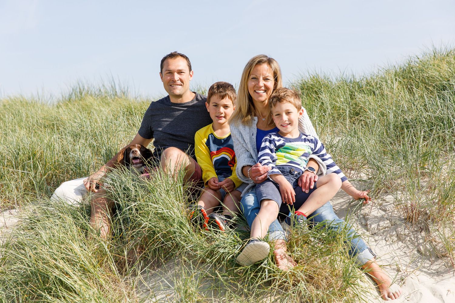A family relaxes together on grassy sand dunes by the ocean during a family beach photoshoot