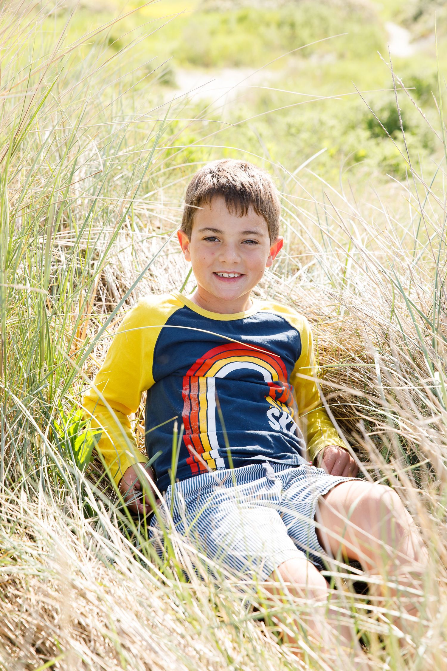 A child wearing a yellow and navy rainbow shirt sits among tall grass on a sunny day during a family beach photoshoot