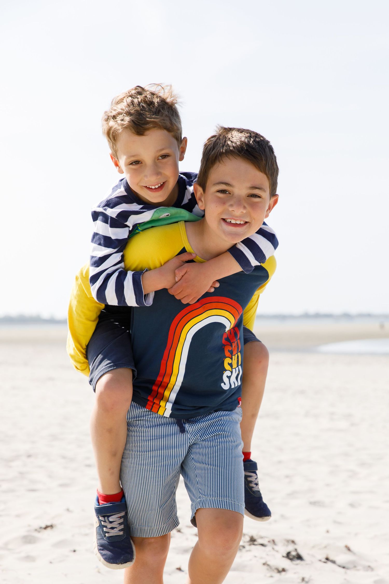 Two children in striped shirts share a playful piggyback ride on the beach during a family beach photoshoot