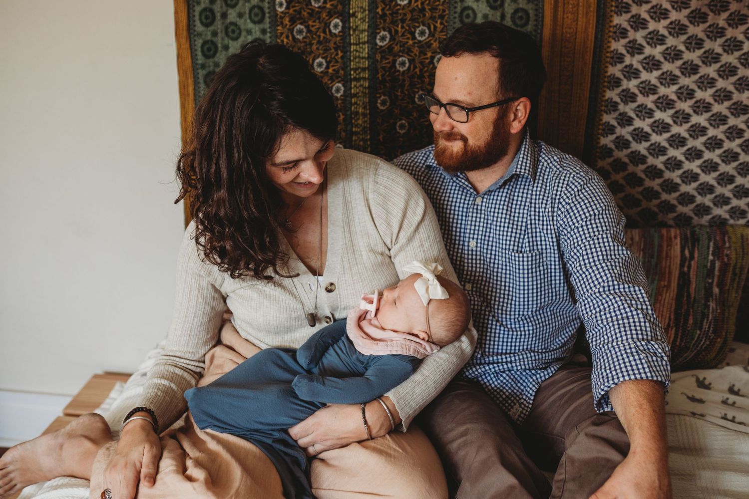 Mom and dad sit on bed holding baby girl with a pacifier while man smiles at his wife