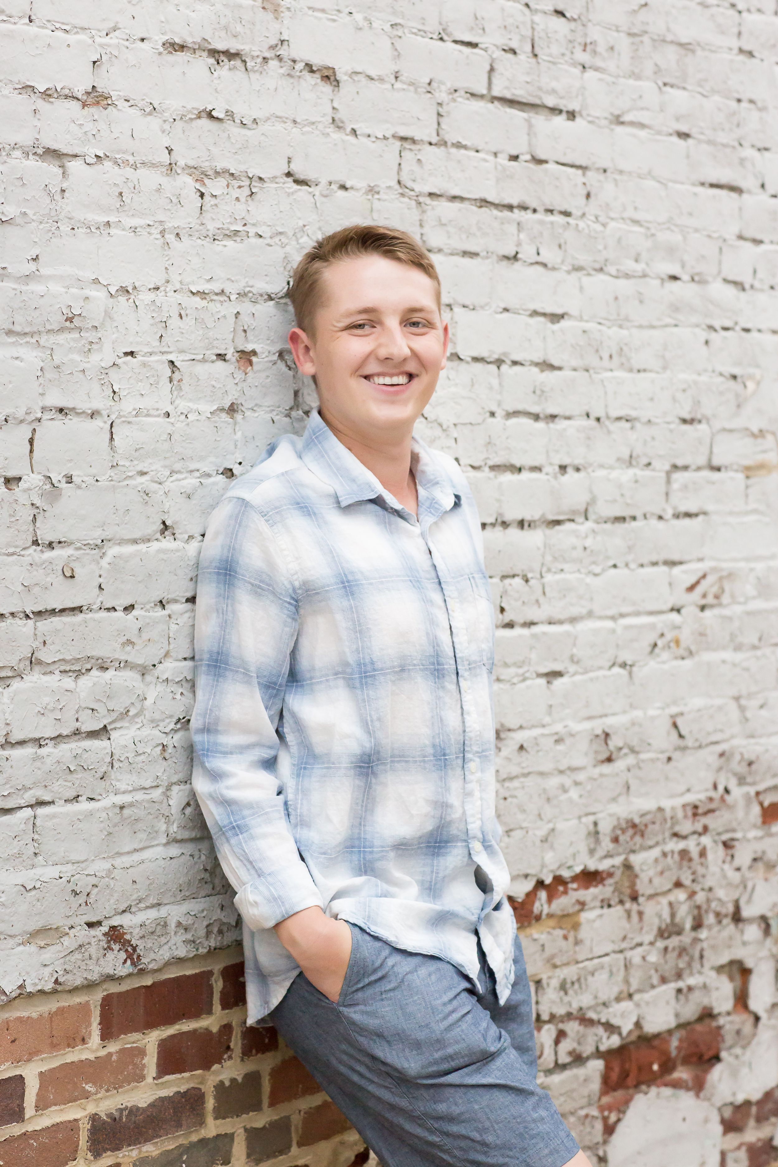 Laughing young man in plaid shirt leans against white painted brick wall with hands in pockets.