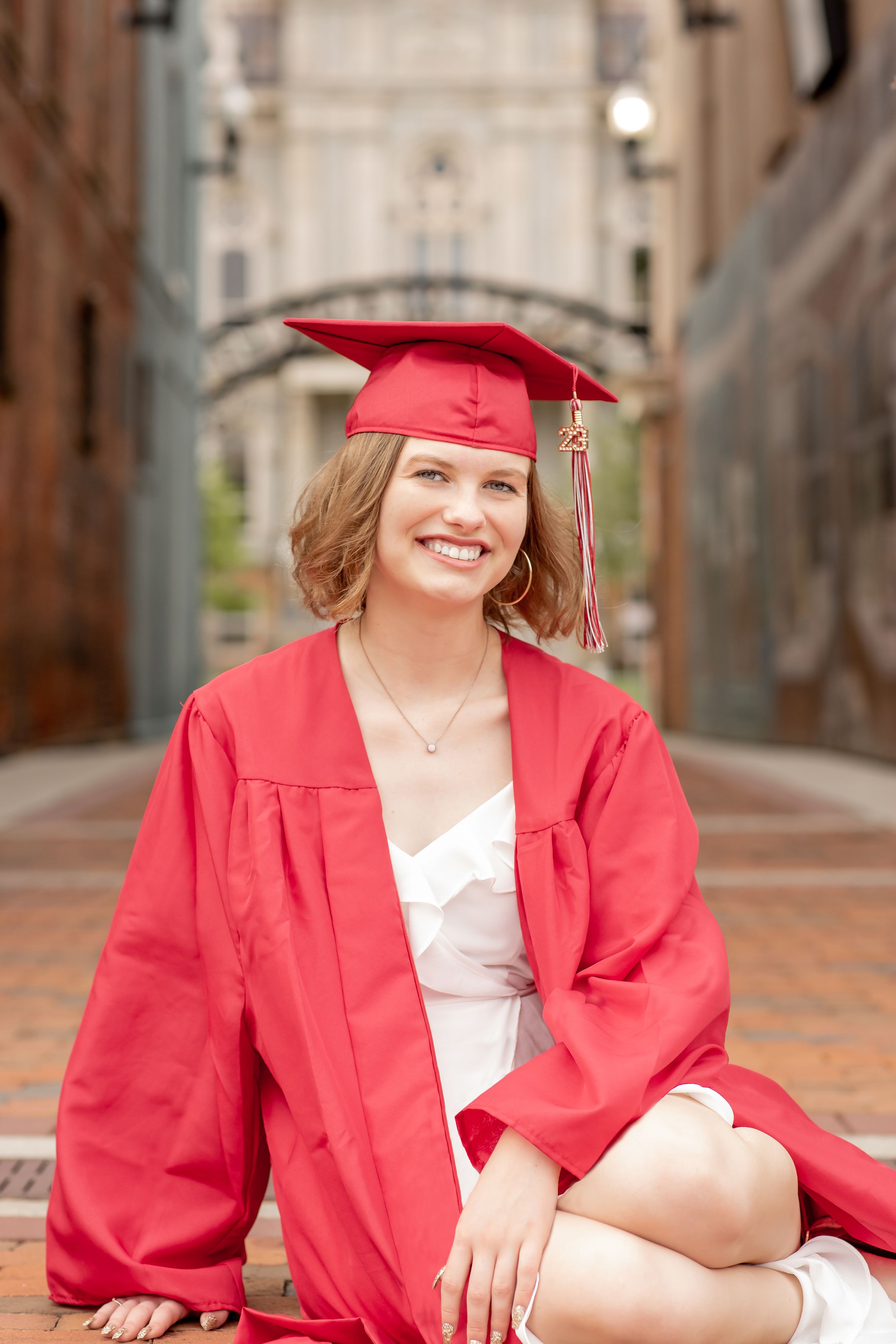 Smiling graduate in red cap and gown sitting on brick path for senior photo.