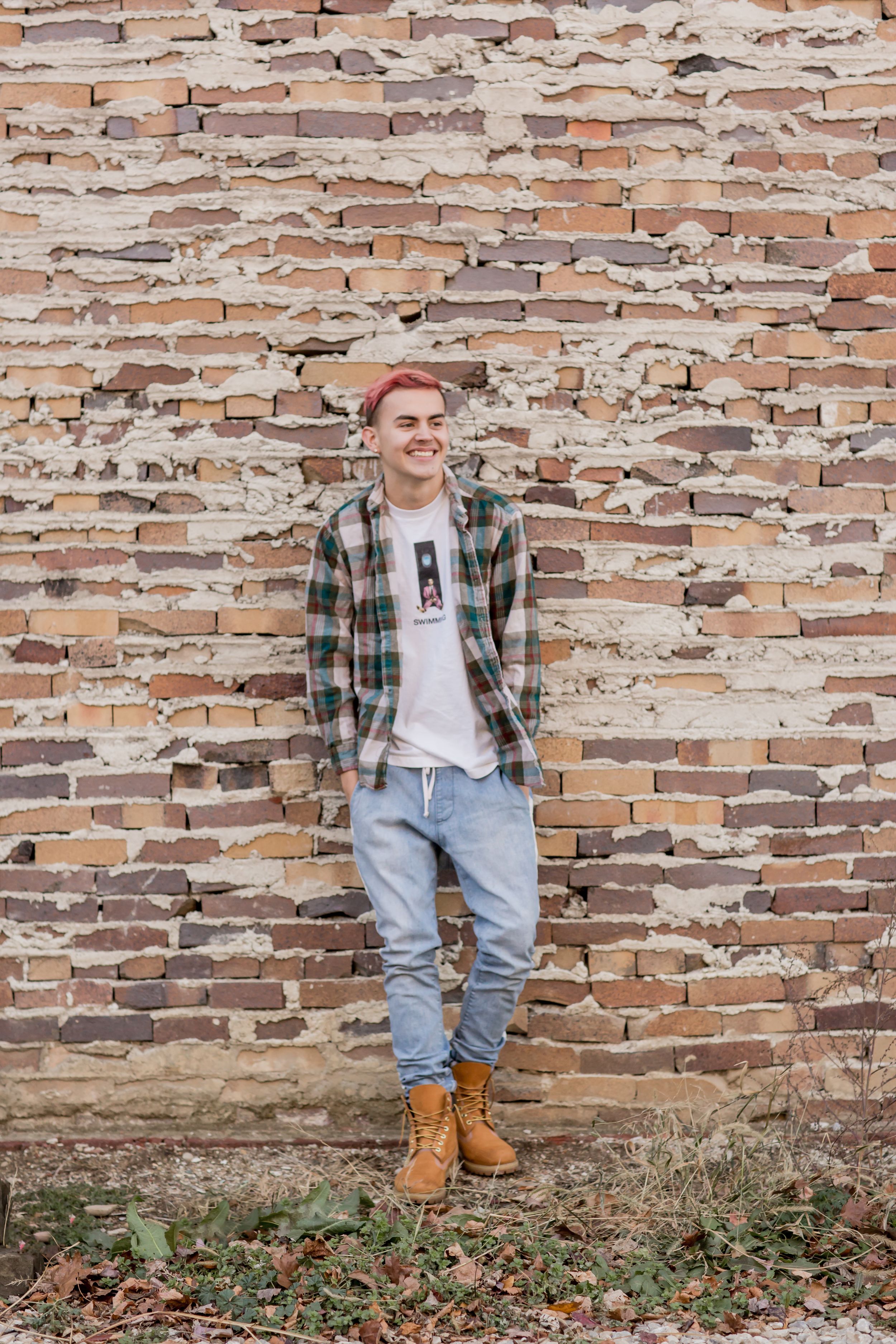 Smiling young man in plaid shirt, jeans, and boots leans against weathered brick wall outdoors.