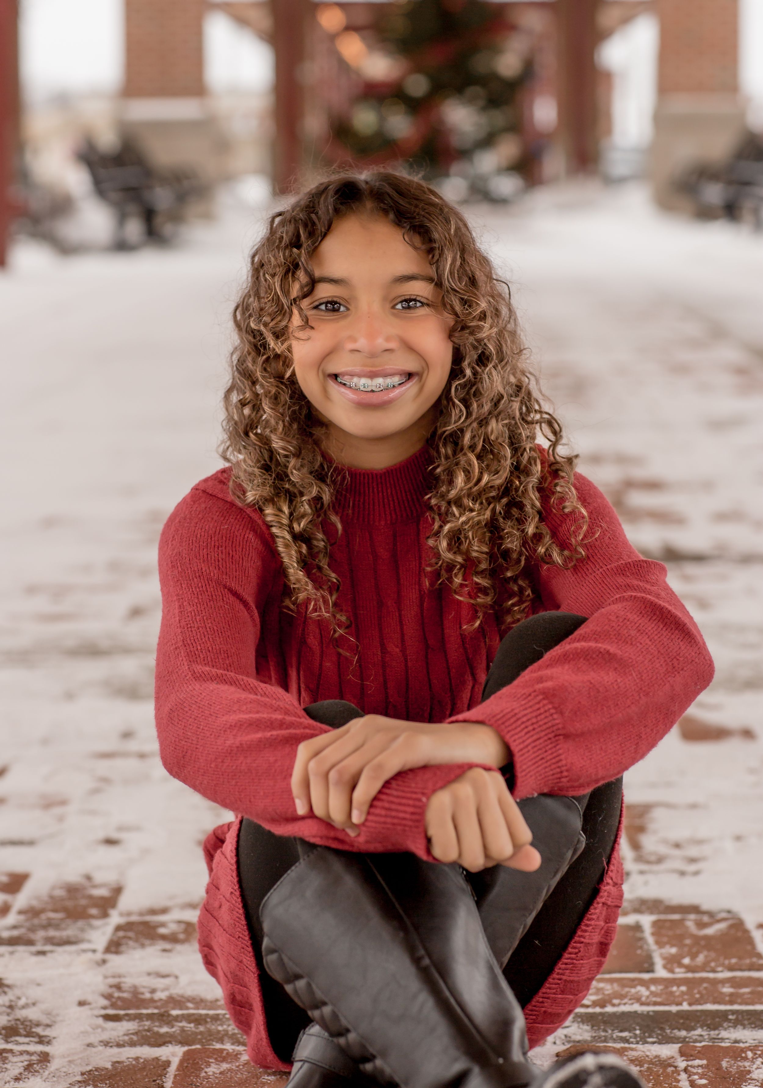 Smiling young girl with curly hair and braces in red sweater sitting outdoors in snow near Christmas tree.