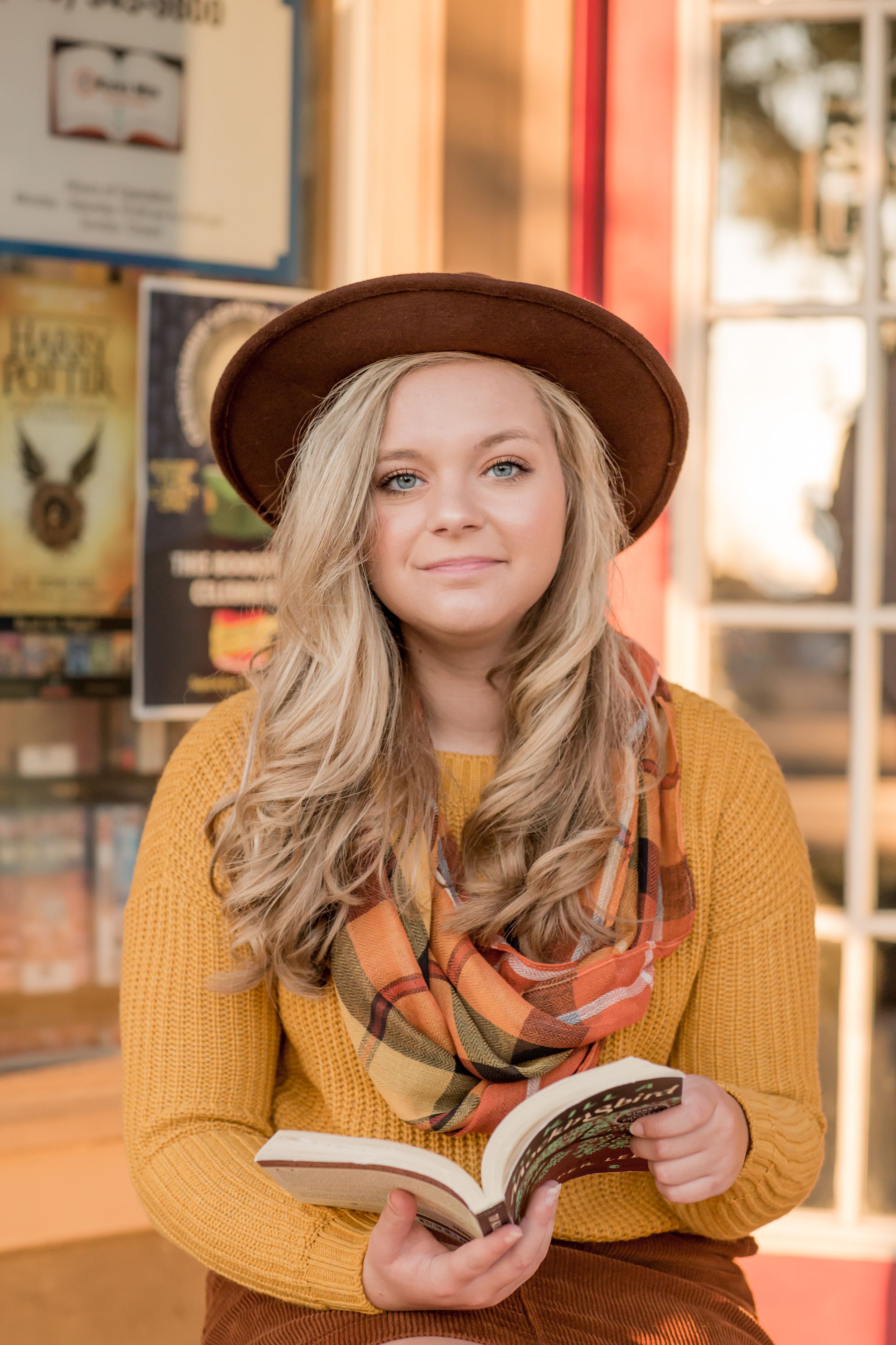 Smiling blonde teen in mustard sweater, brown hat, and plaid scarf holds open book outside bookstore.