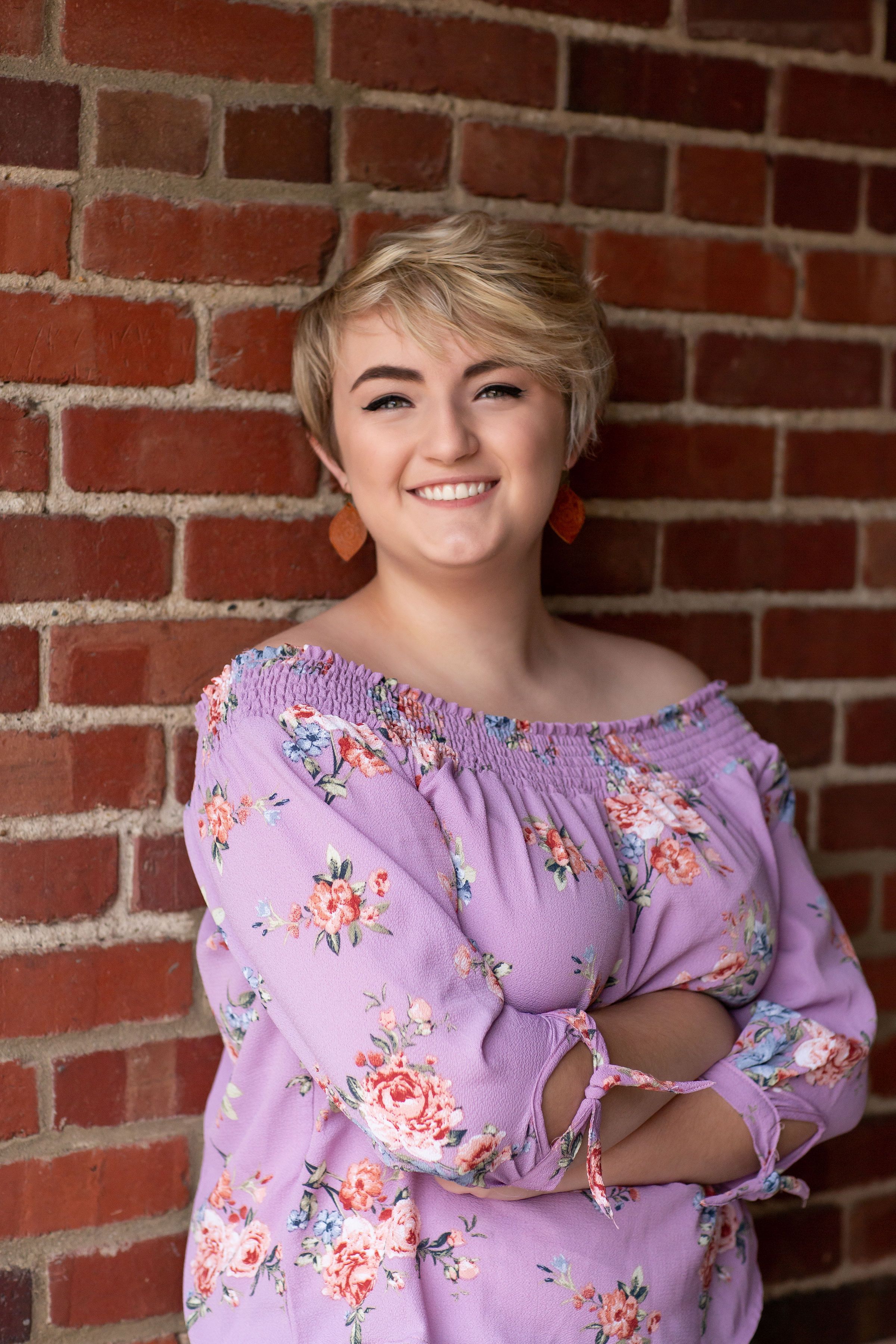 Smiling young woman with short blonde hair in floral off-shoulder lavender top poses against red brick wall.