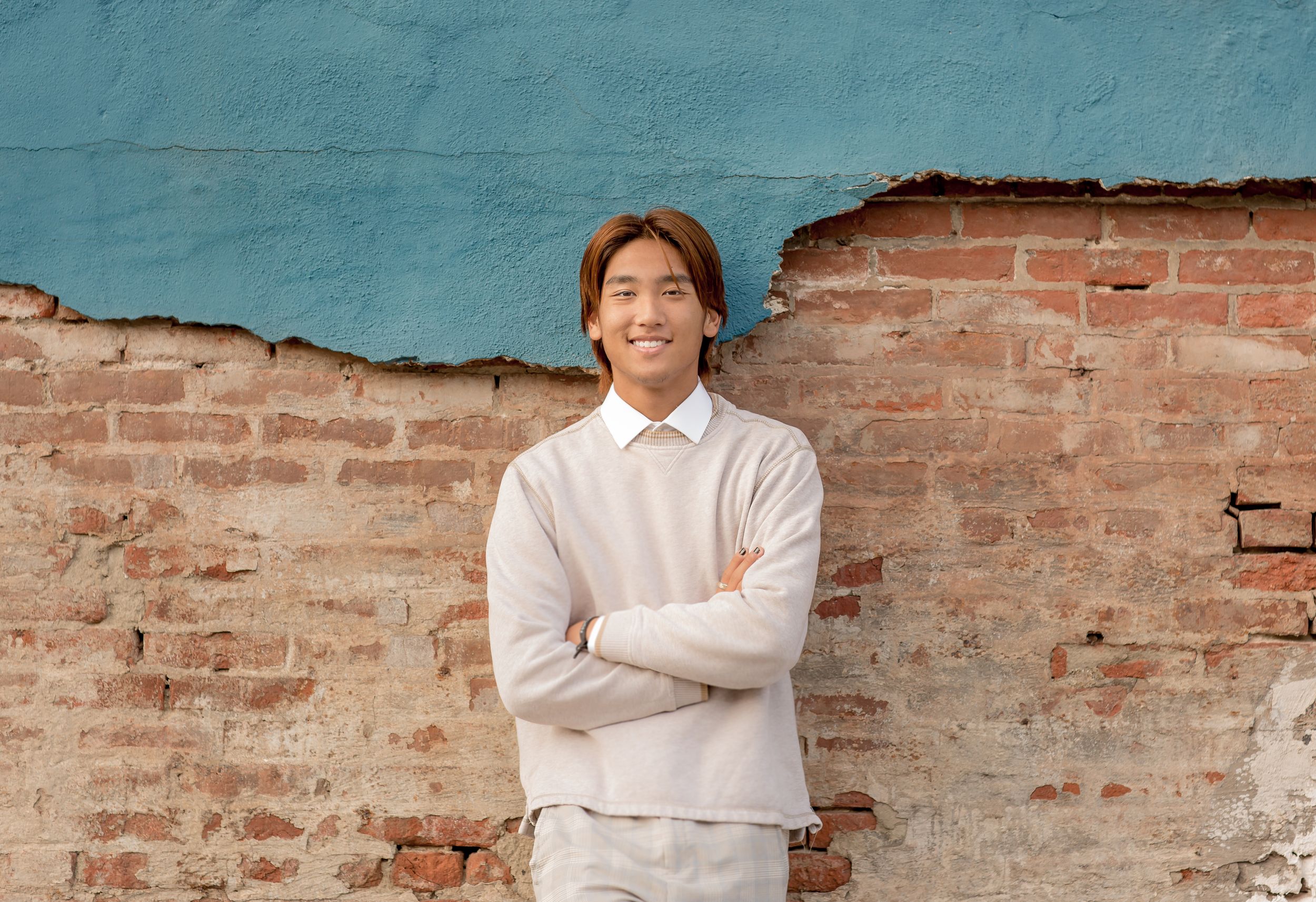 Young man in beige sweater stands with arms crossed smiling before weathered turquoise brick wall.