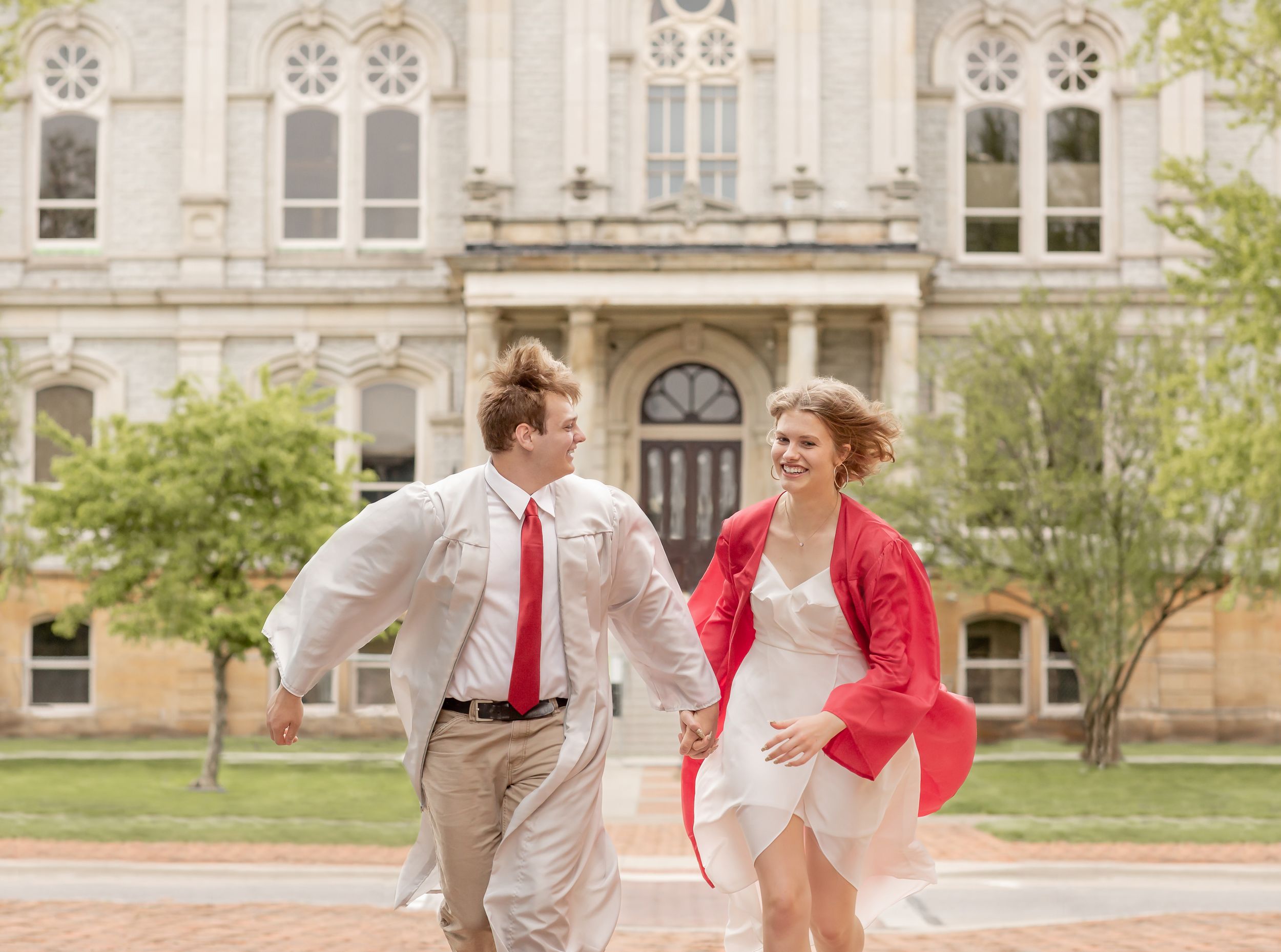 Two graduates in red and white running and laughing in front of historic building.