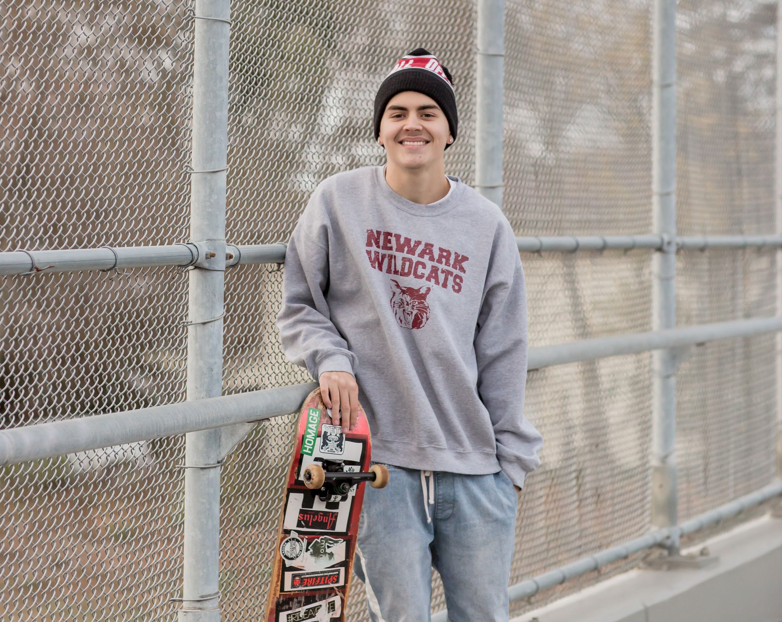 Smiling teen boy in Newark Wildcats sweatshirt and beanie holds skateboard at outdoor skate park.