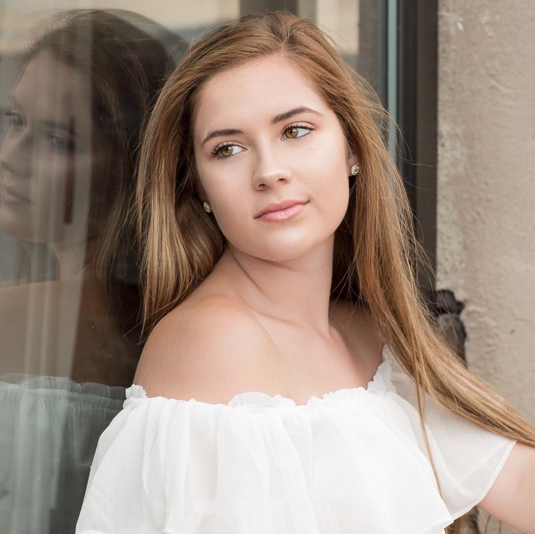 Young woman with long brown hair in white off-shoulder top posing against glass window outdoors.