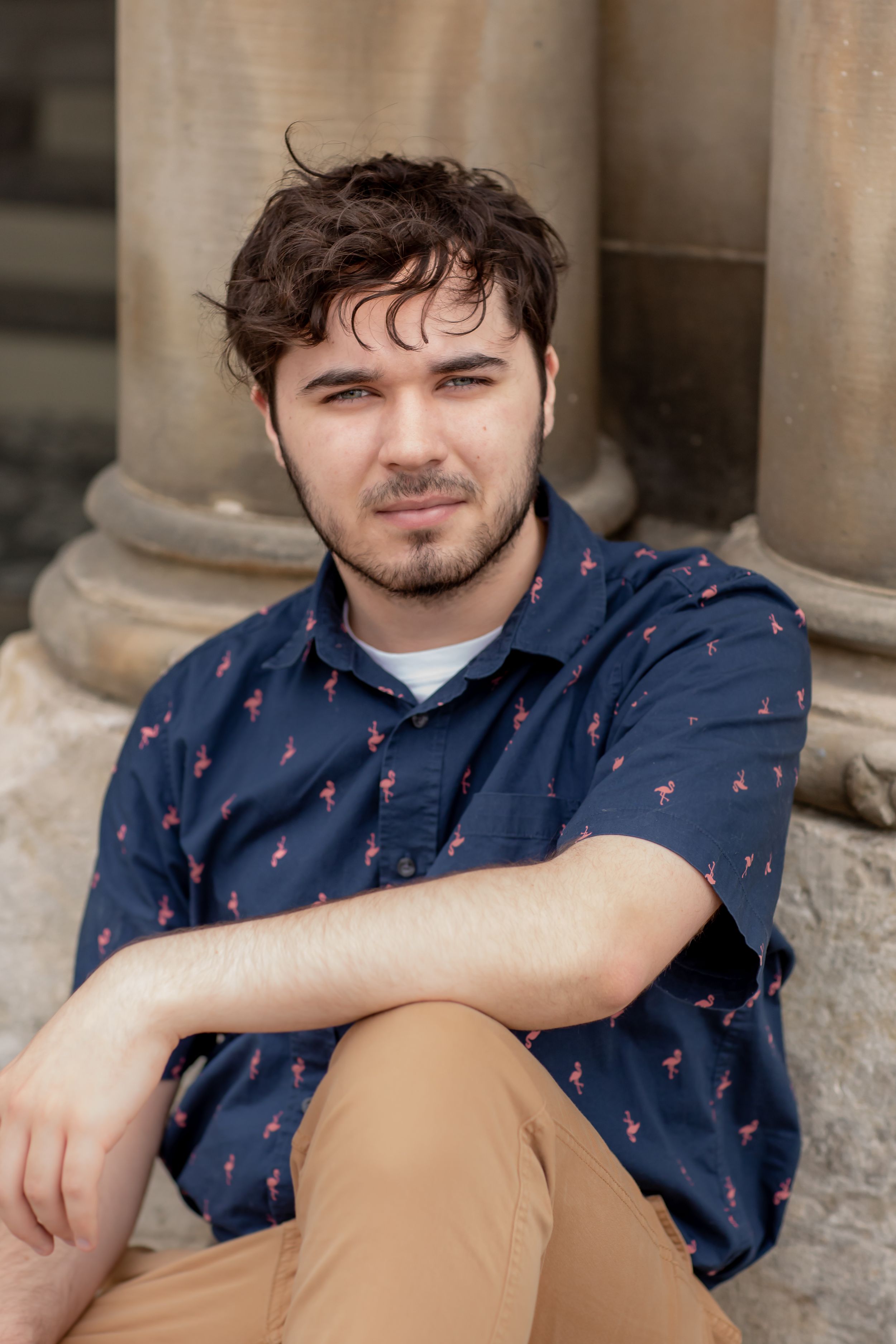 Young man in flamingo print navy shirt sitting on steps near stone columns.