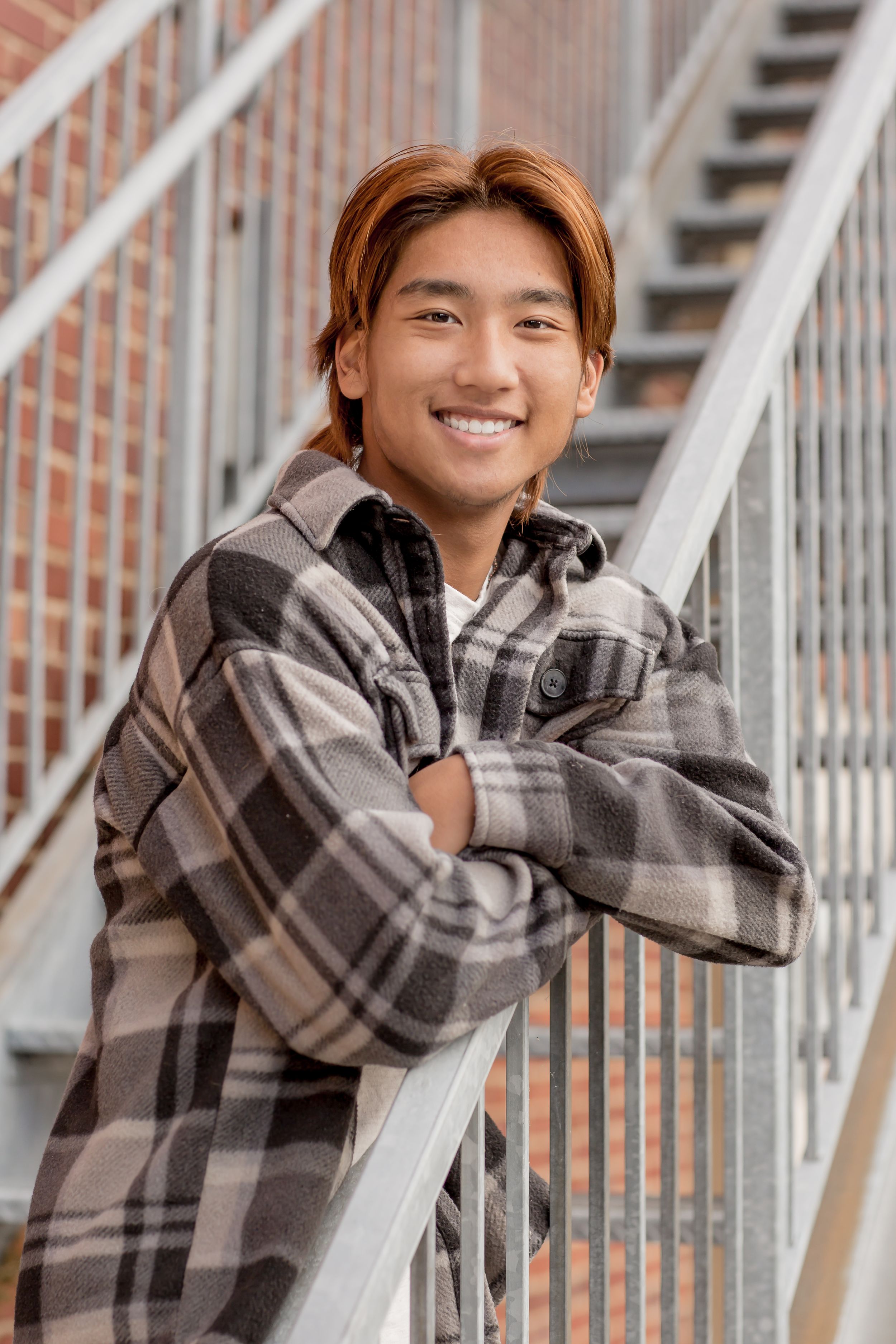 Smiling young Asian man with auburn hair in plaid shirt leaning on metal staircase railing outdoors.