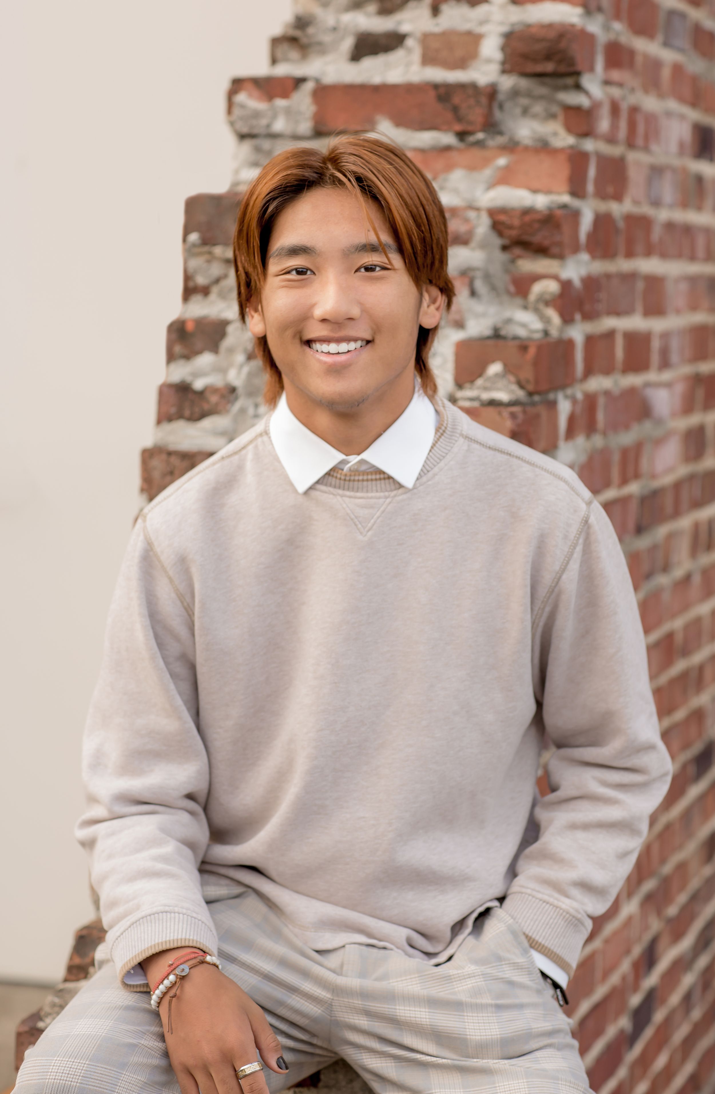 Smiling young man with auburn hair in beige sweater sits against exposed brick wall for portrait.