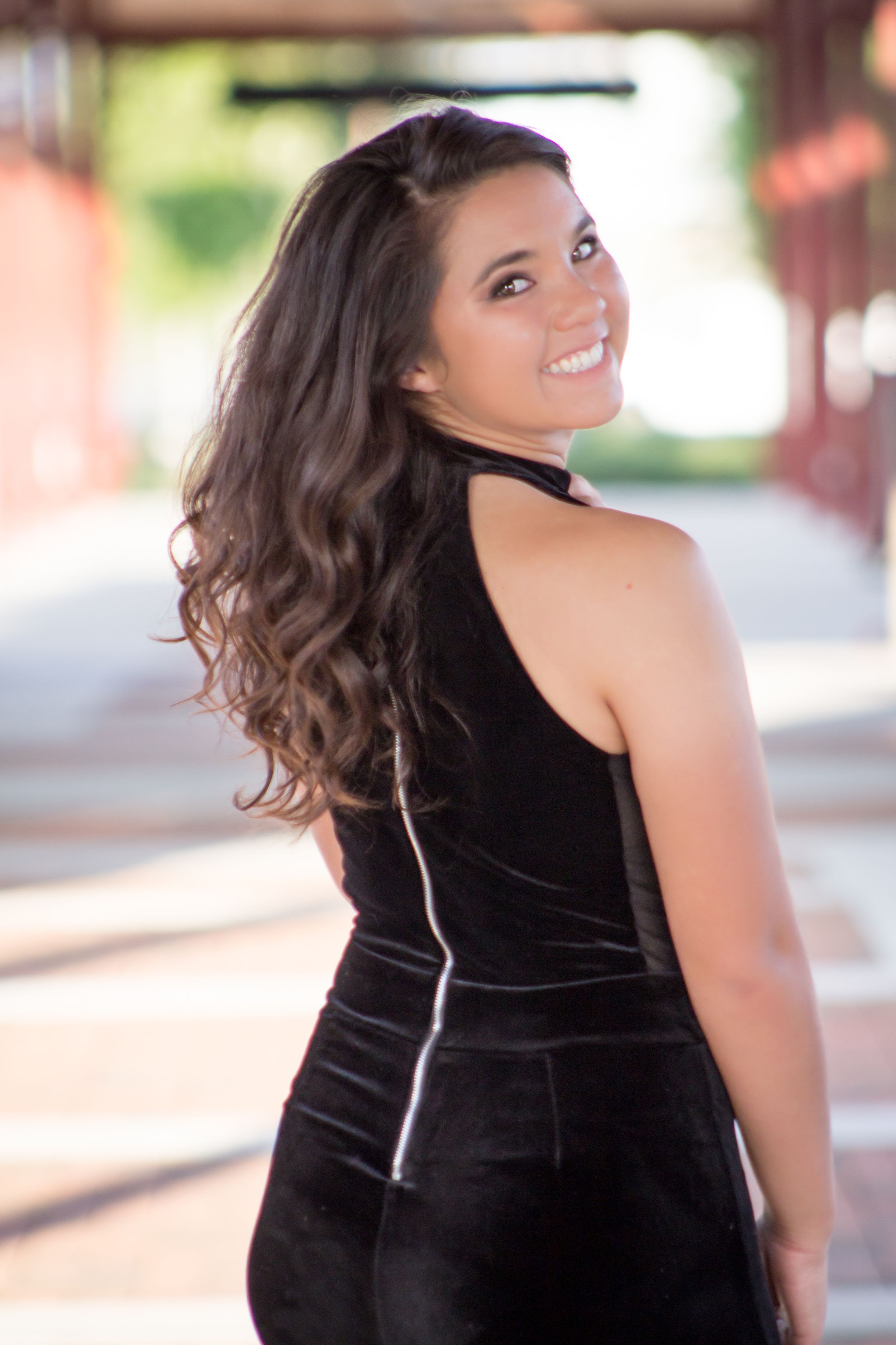 Brunette woman in black velvet outfit smiling over shoulder for senior portrait.