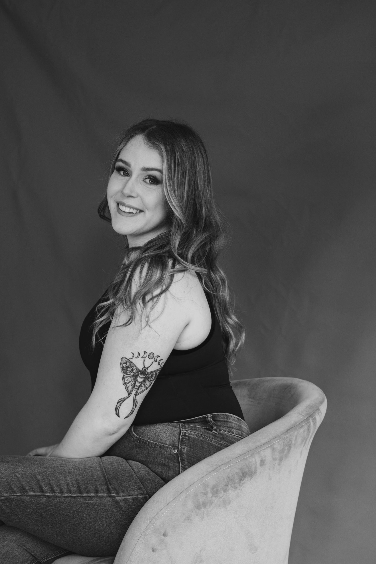 Portrait of someone sitting in velvet chair showing off floral tattoo in black and white.