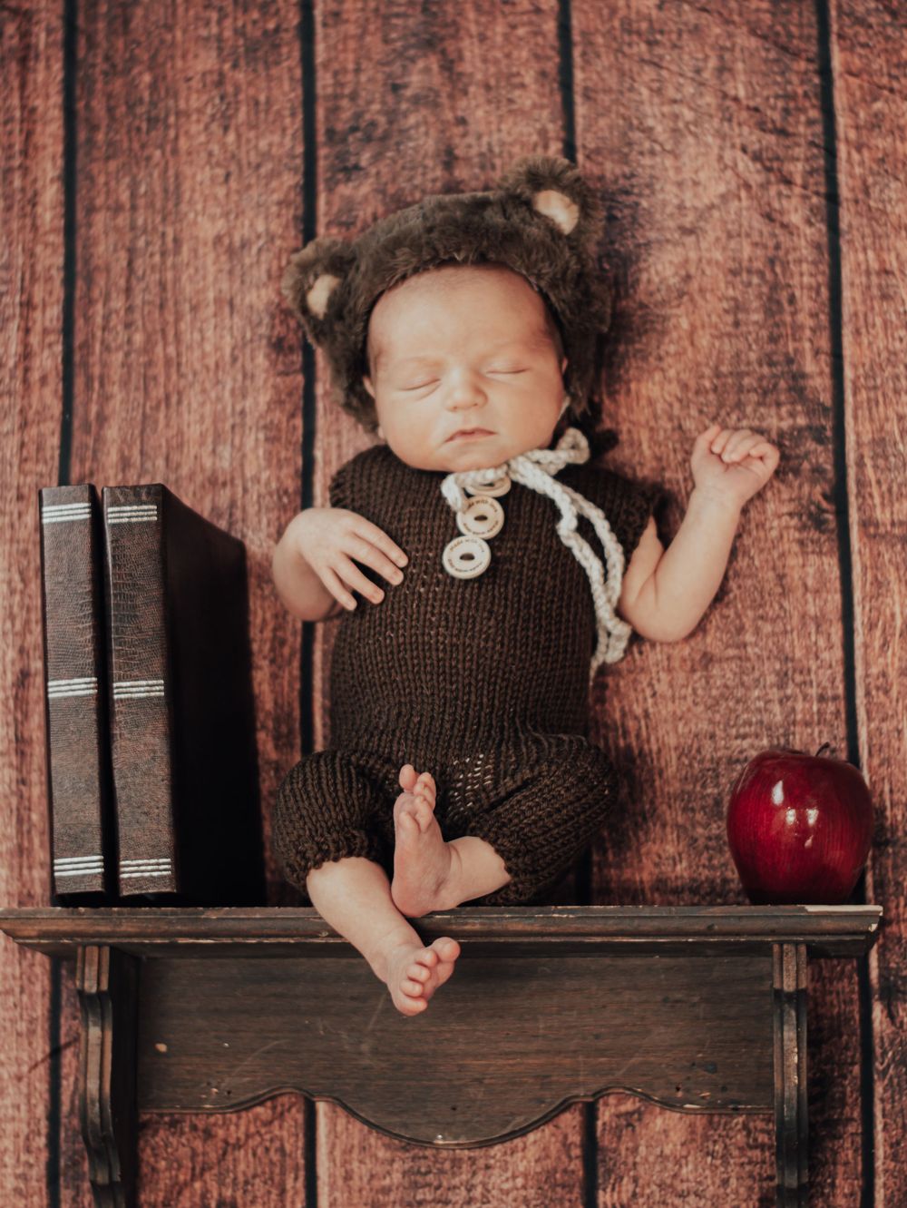 Sleeping newborn in brown knit outfit with teddy bear hat lies on rustic wooden shelf with books and red apple.