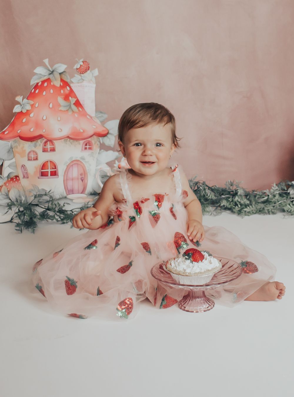 Baby in strawberry dress sits by whimsical mushroom houses with pink dessert.