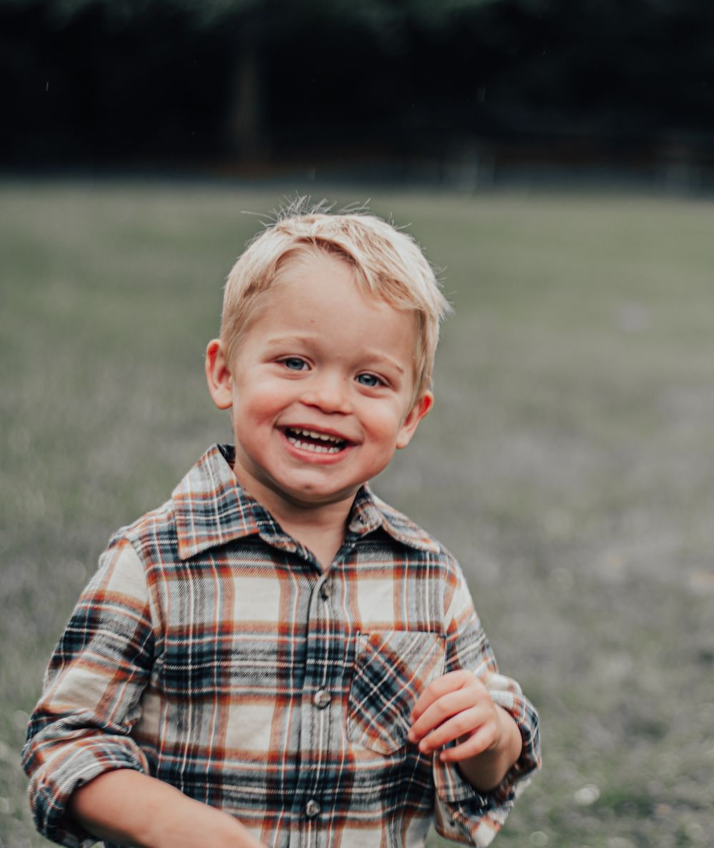 Young child in plaid flannel shirt smiling outdoors in grassy field.