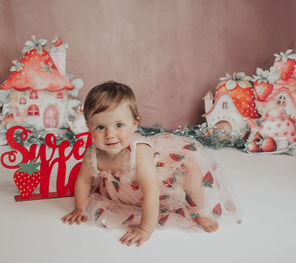 Baby in pink strawberry dress crawling on white floor with mushroom house decorations in background.