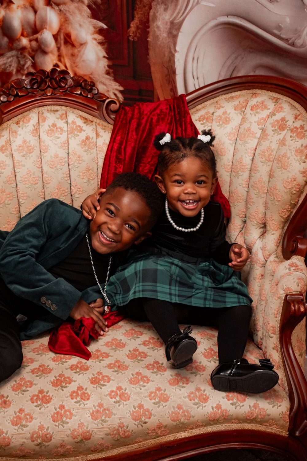 Two children sharing joyful moment on vintage floral couch with red and pink holiday decorations.