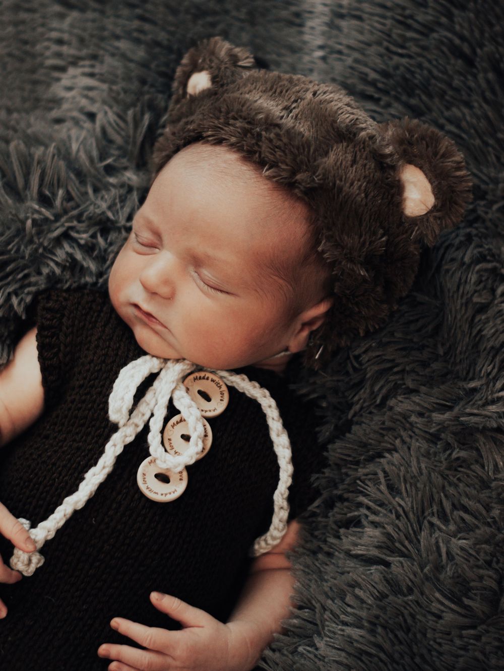 Peaceful sleeping baby wearing brown knit outfit and bear ear hat on dark furry blanket.