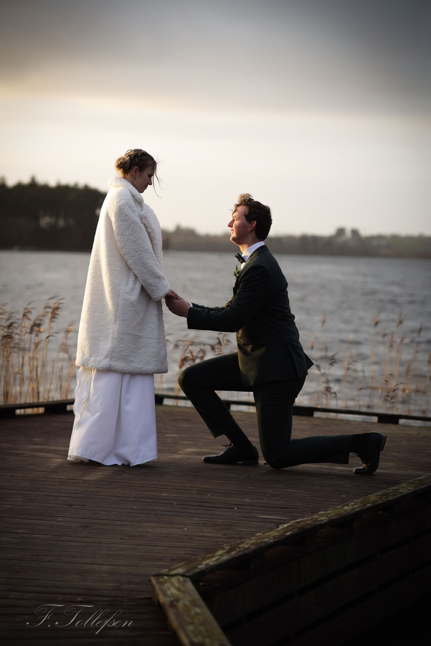 Groom kneeling on one knee holding brides hands on a lakeside dock at golden hour sunset.