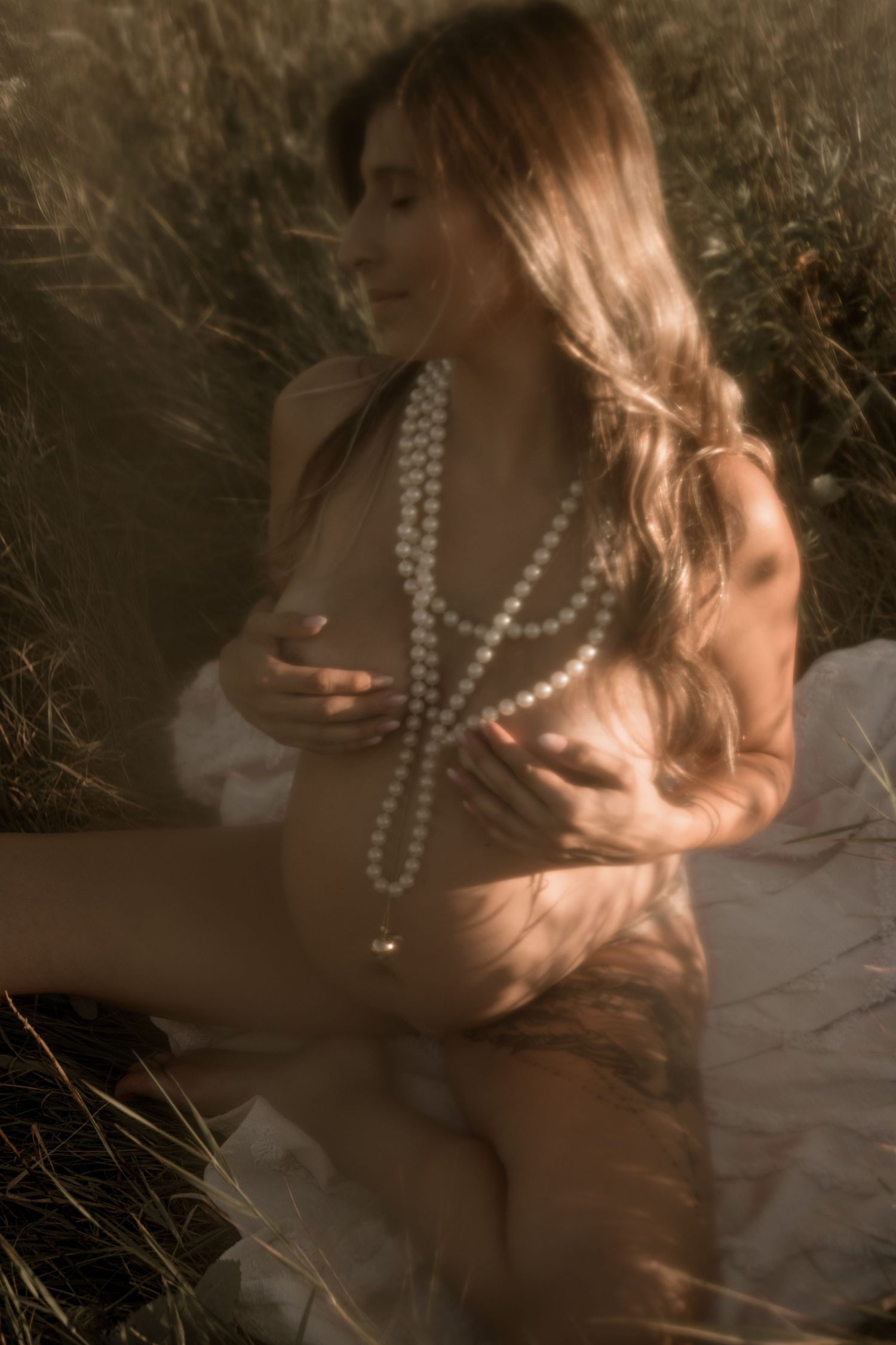 Artistic sepia-toned boudoir photo of someone wearing pearl necklaces in soft natural light among tall grasses.