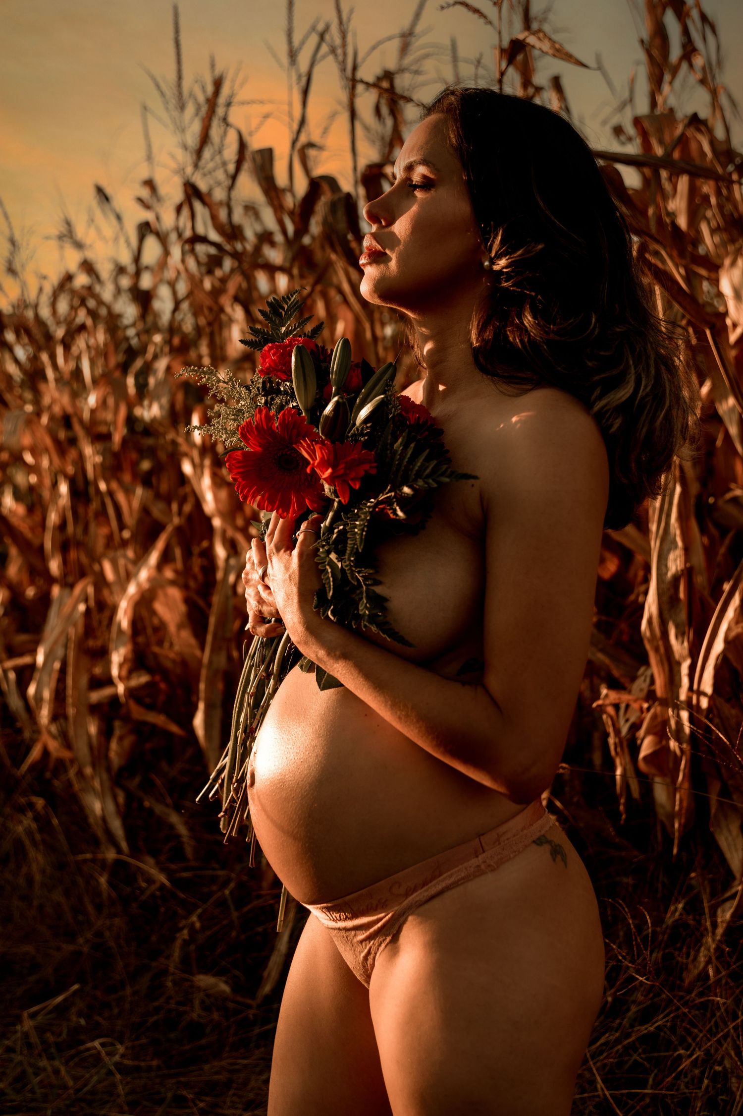 Artistic maternity photo in a cornfield at sunset with red roses creating a warm, intimate mood.