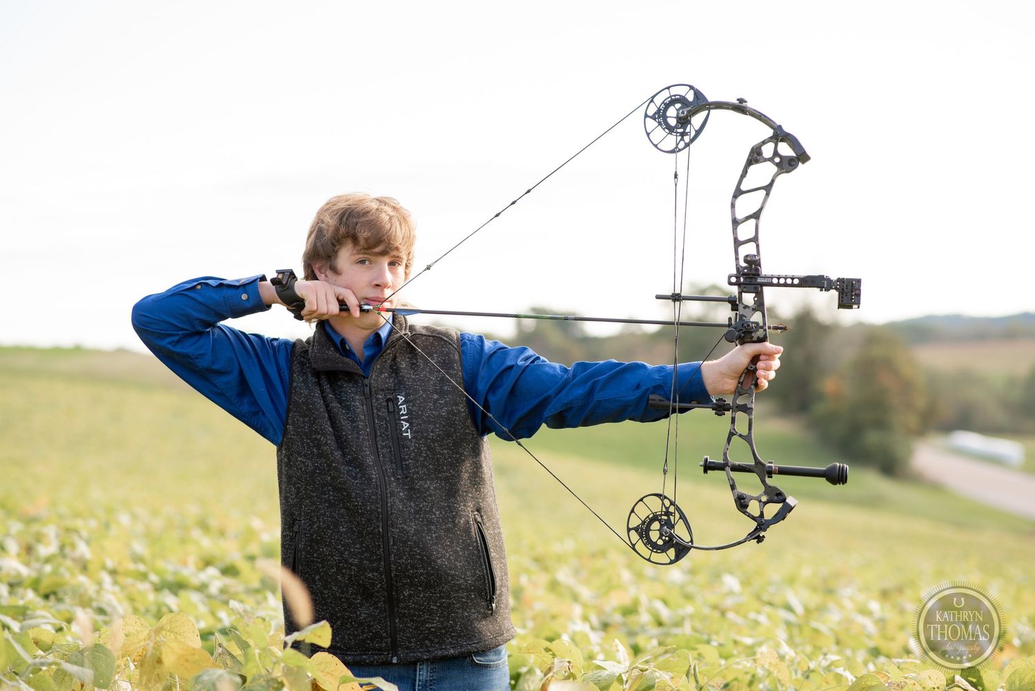 An archer draws back a compound bow while wearing a blue and gray vest outdoors.