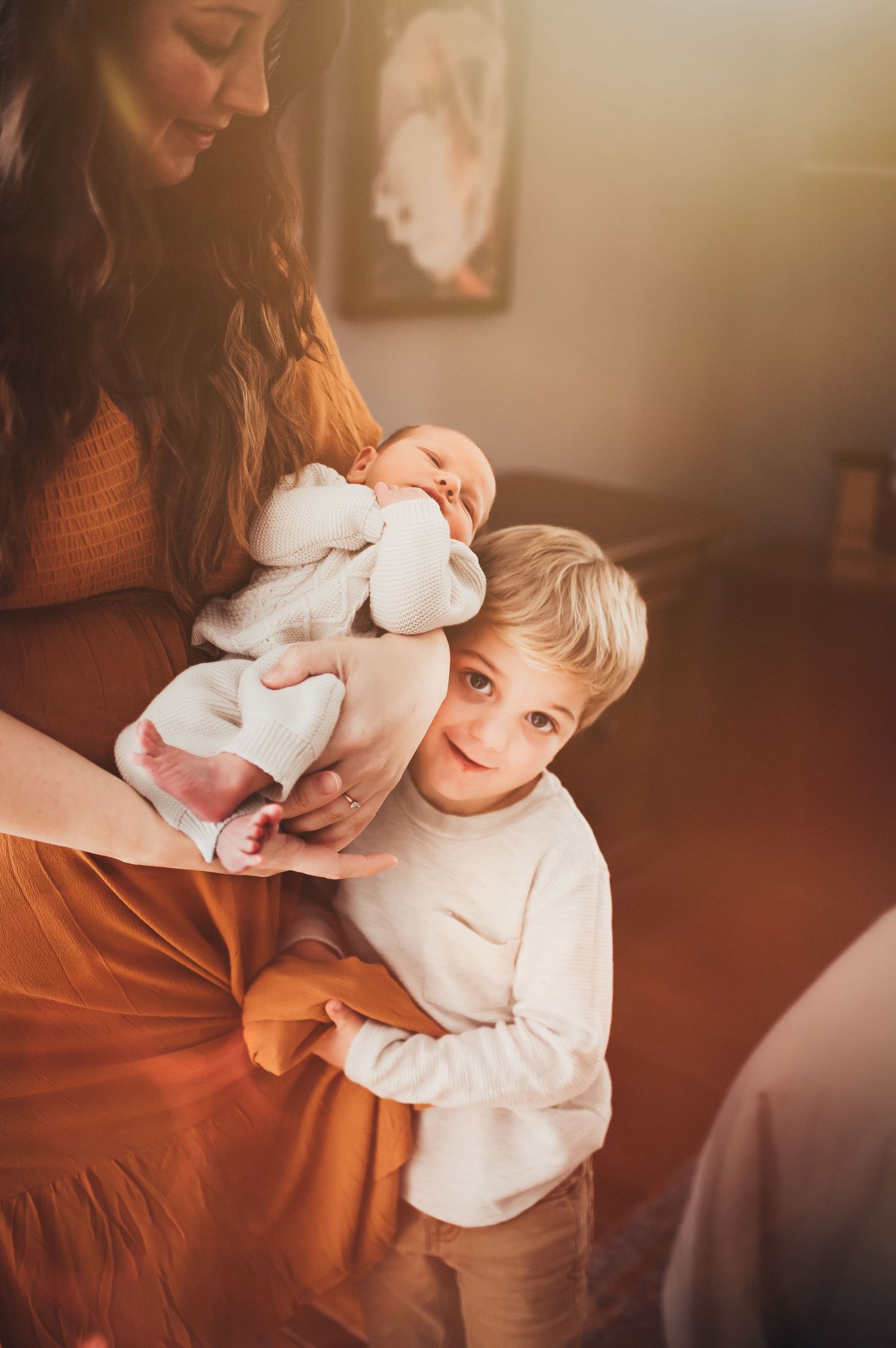 big brother wraps himself in his mom's skirts as she holds newborn little brother at lifestyle newborn session
