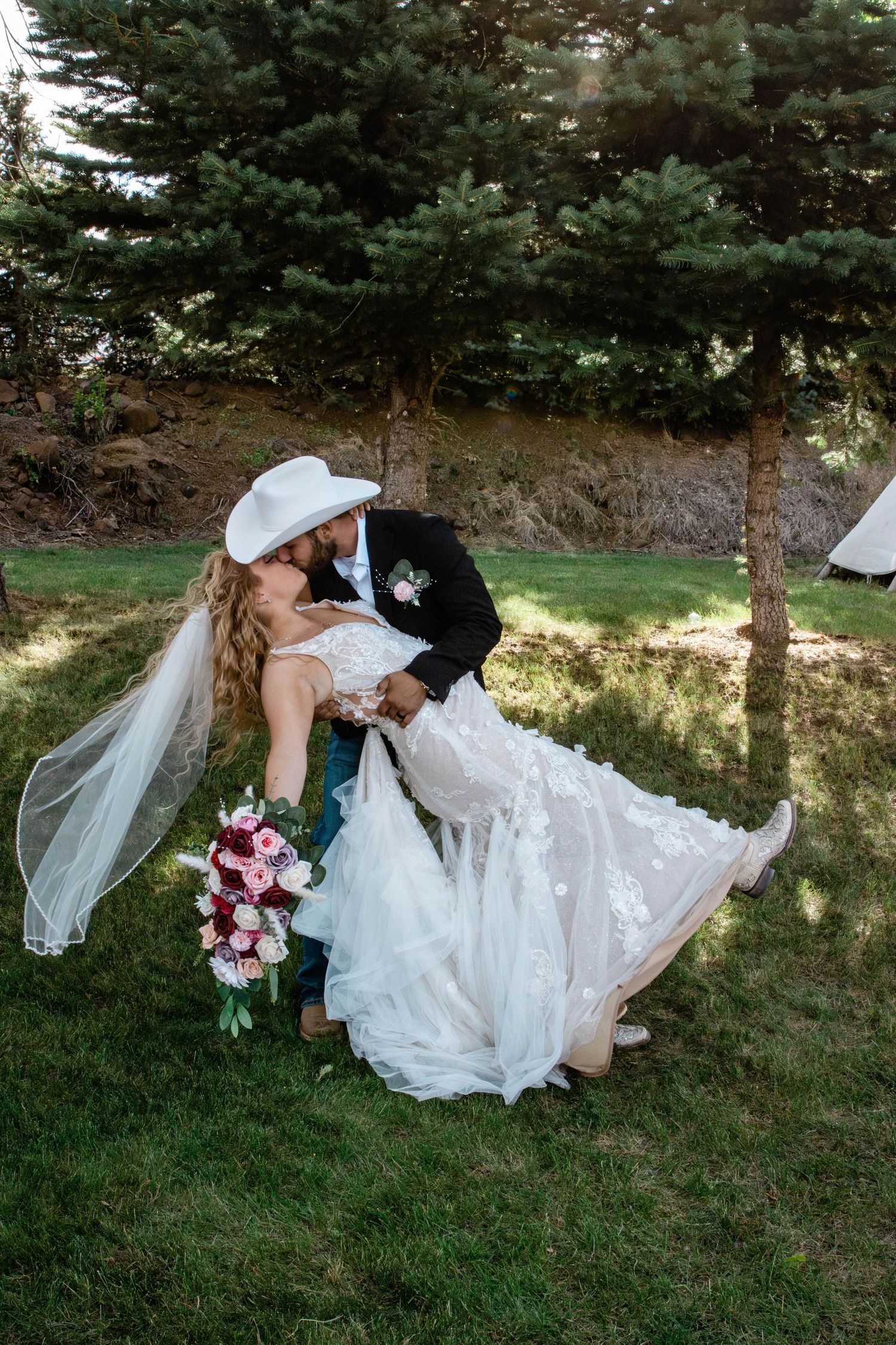 Groom in cowboy hat dips bride in floral wedding gown for a romantic kiss outdoors.