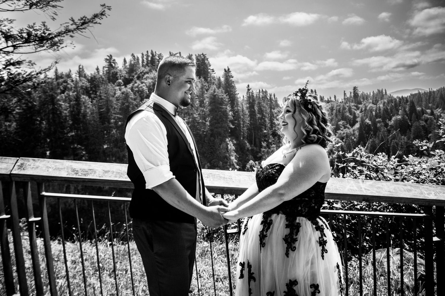 Black and white photo of couple holding hands on scenic overlook with forest backdrop.