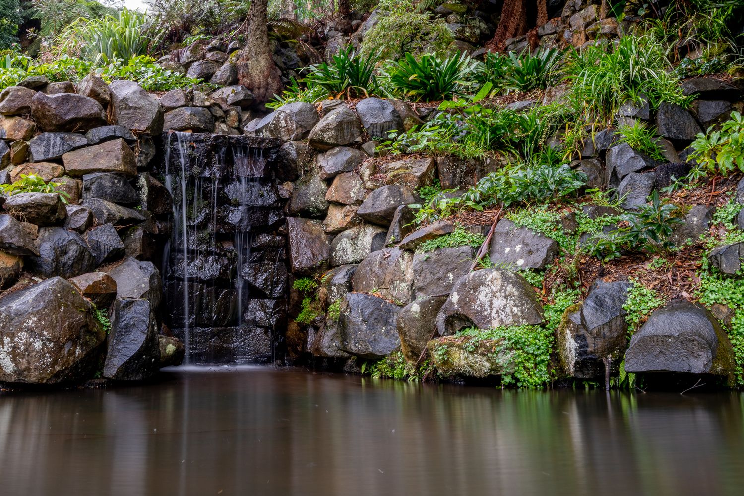Small waterfall cascading over mossy rocks into a still pond surrounded by lush green garden plants.