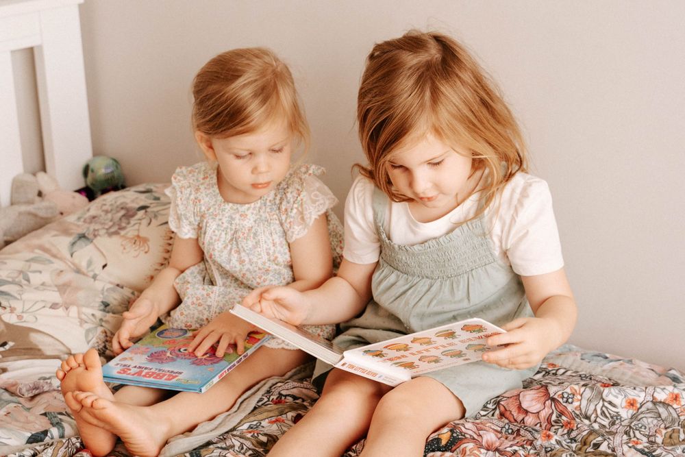 Two children reading a book together on a floral patterned bed.