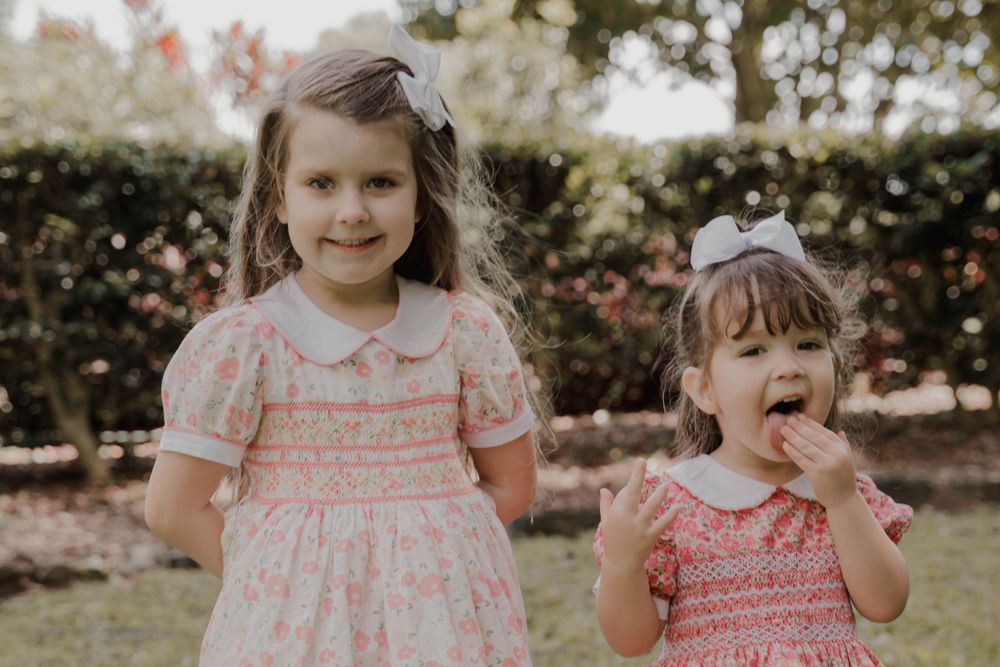 Two little girls in vintage-style dresses pose together in a garden.