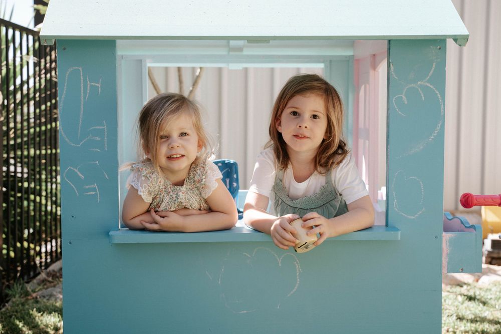 Two children playing in a turquoise playhouse window in a sunny backyard.