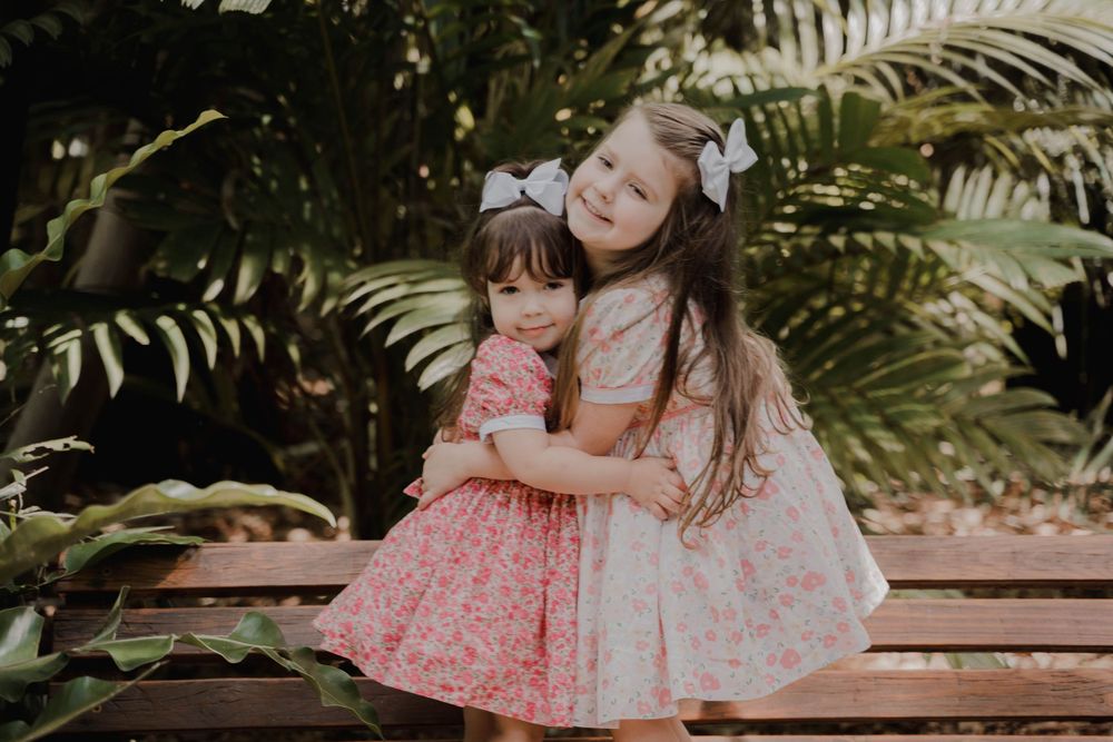 Two children in matching pink dresses share a sweet embrace on a wooden bench.