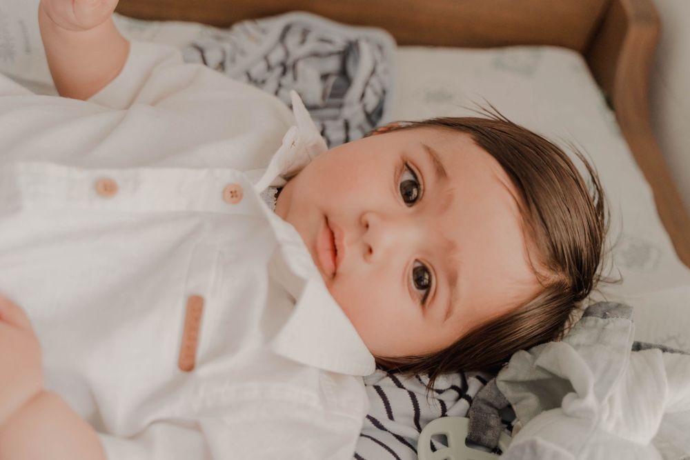 A baby lying on white bedding looks directly at the camera with wide eyes.